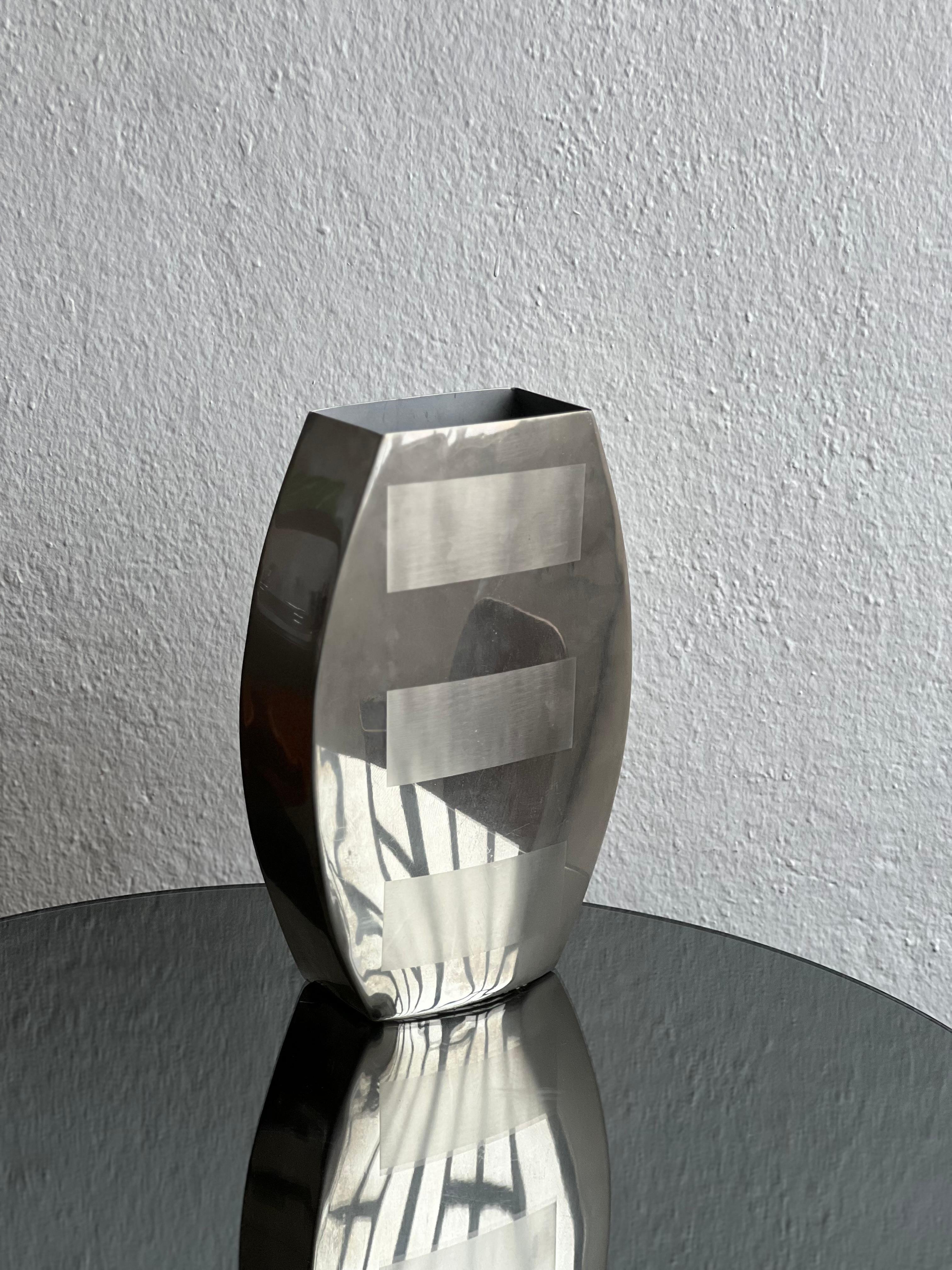 Este jarrón vintage de los años 70 emana una estética elegante y modernista típica de la sensibilidad de diseño de la época. Fabricada en acero inoxidable pulido, su superficie brilla con un acabado de espejo que refleja la luz y el entorno con