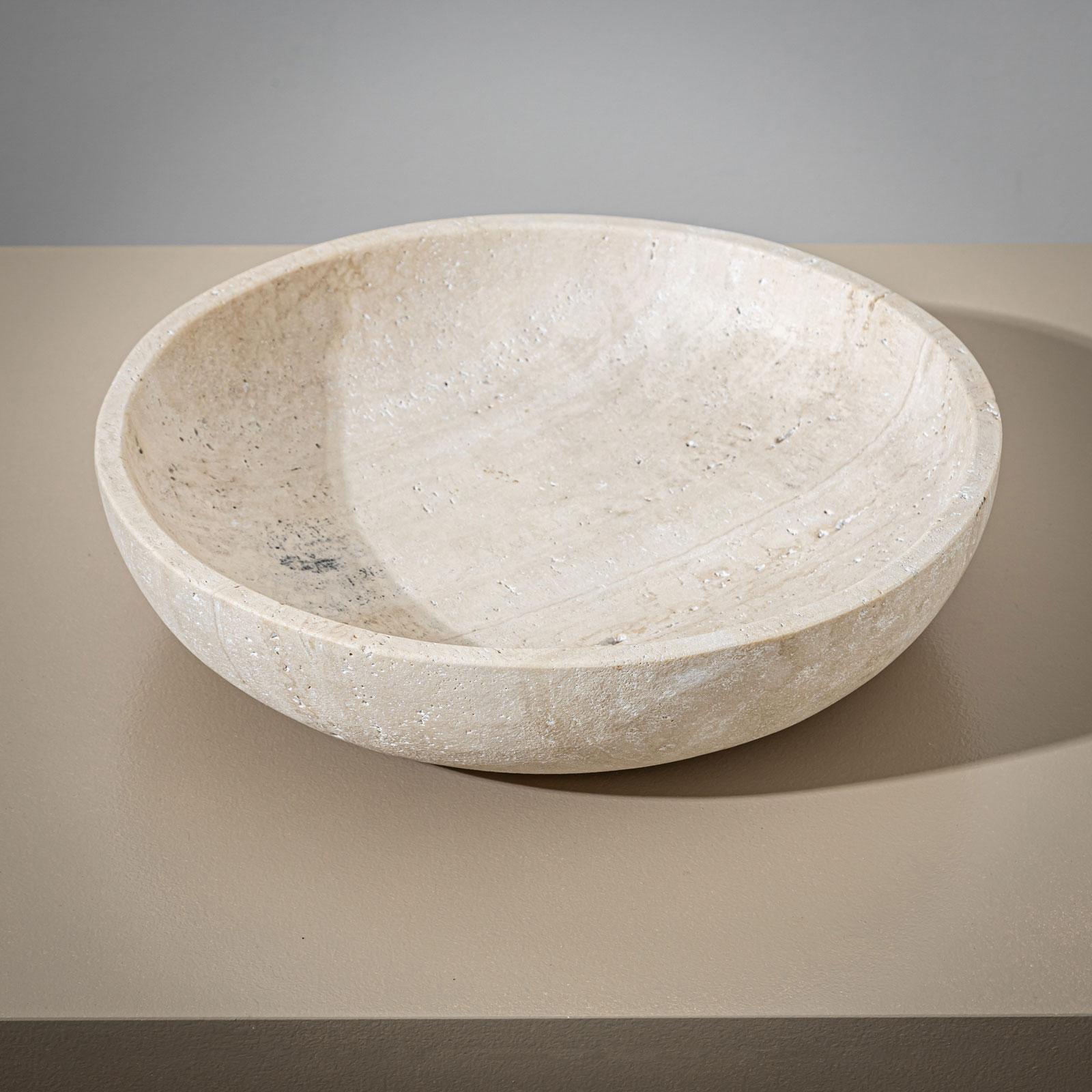 A large, heavy stone bowl crafted from beige travertine, featuring a minimalist design. The bowl is distinguished by its high rim and flat shape, exuding simple yet timeless elegance.