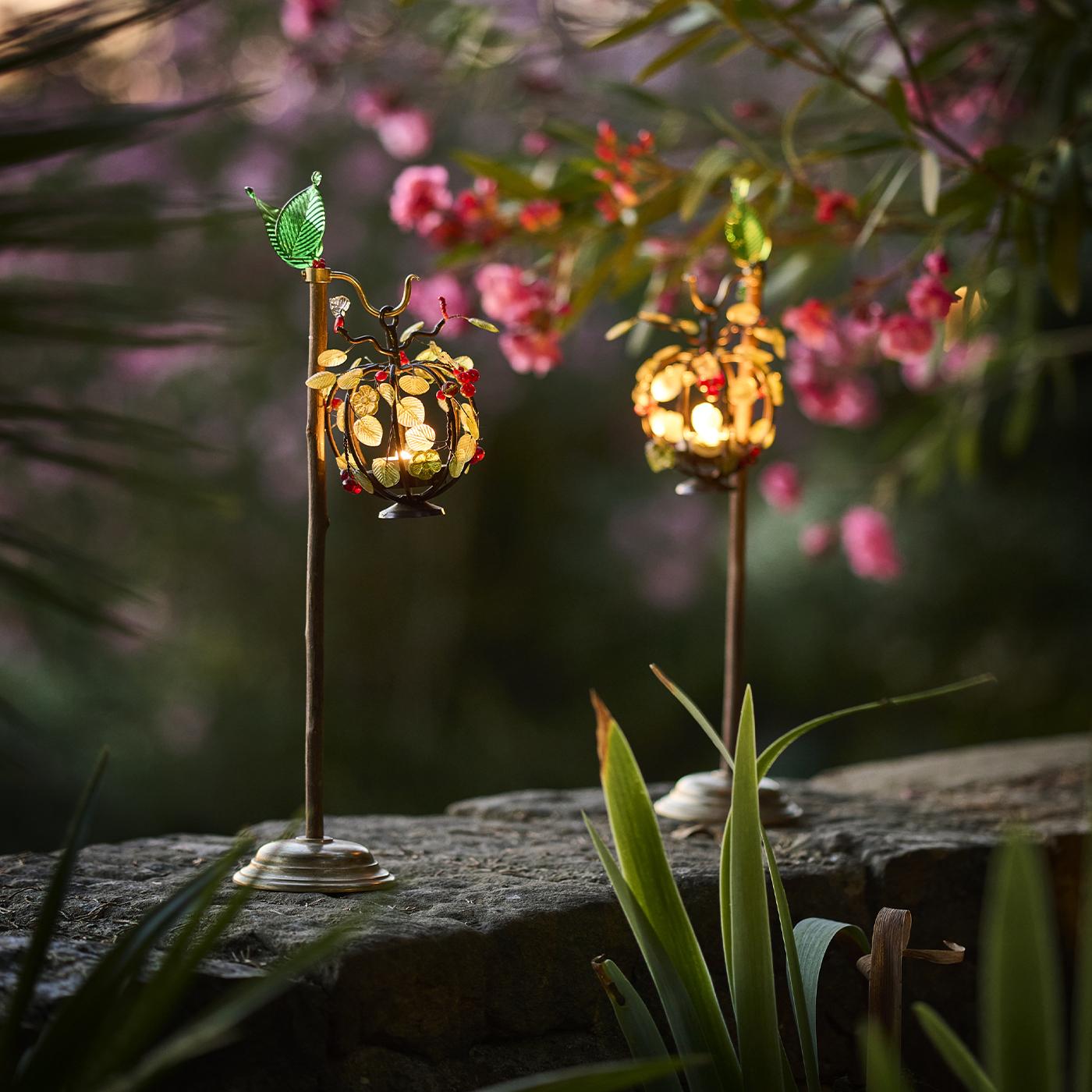 Dieser dekorative Kerzenhalter aus Holz besteht aus einem schlanken Holzstiel mit einem Sockel und einem Halter aus Messing, der die Kerze umschließt. Die Kerze ist von fein gearbeiteten Metallblättern und Glasbeeren umgeben, die den optischen Reiz