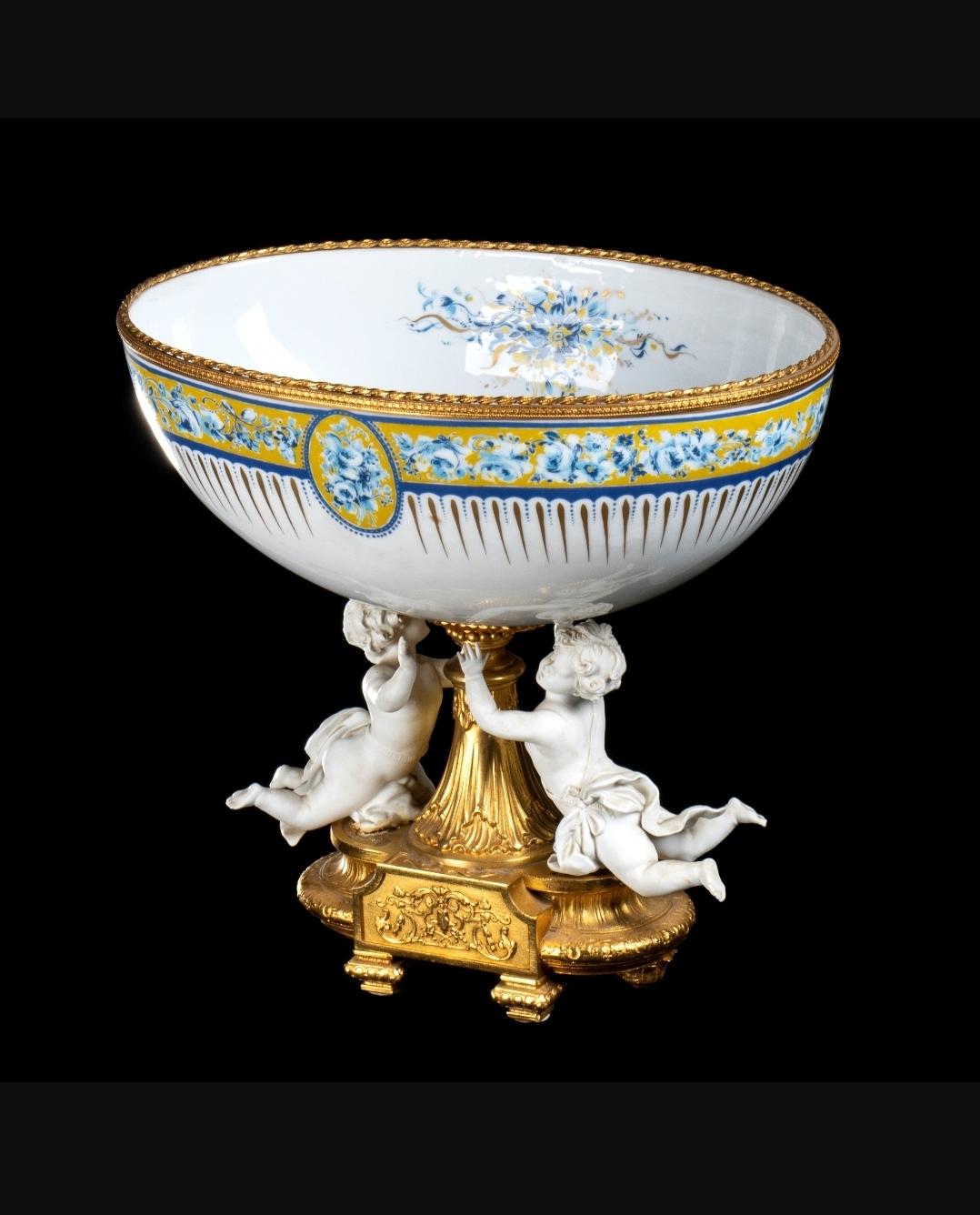 Decortiche marcate (Tiche, Porcellane d'Arte). Magnifico centrotavola in stile romano con ciotola ovale con bordo floreale blu e bordo in metallo dorato su una base in porcellana di bisquit e metallo dorato. MISURE: 15″ di larghezza x 14-1/2″ di