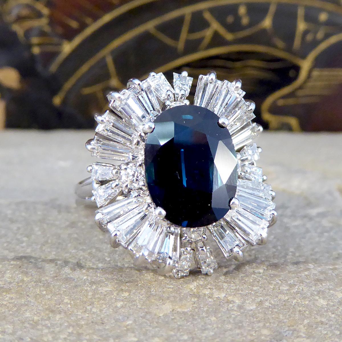 Presenting our Sapphire and Diamond Ballerina cluster ring, a pinnacle of sophistication. This ring boasts a 4.33ct oval cut Sapphire showing a deep and dark blue hue, enveloped by tapered baguette diamonds weighing a total of approximately 2.20ct