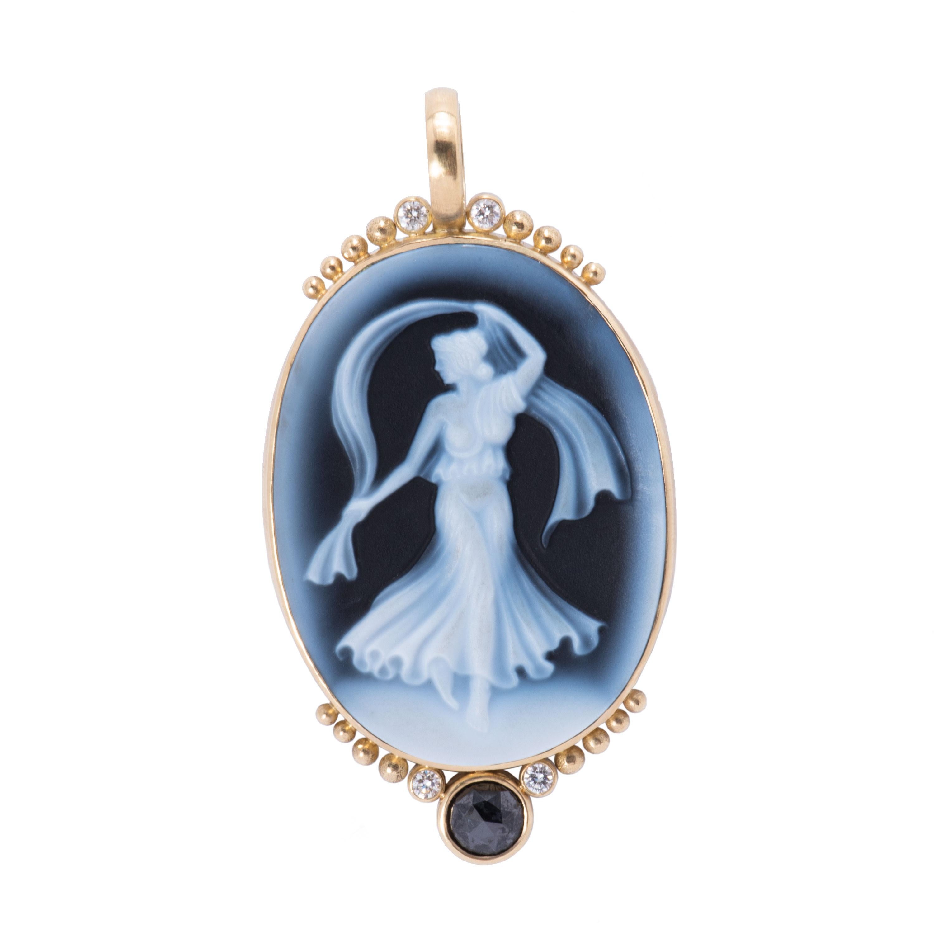 A large blue cameo pendant in carved agate depicts a woman dancing with a scarf in a diaphanous gown against a deep blue. Framed in 18 karat gold, the pendant is embellished with gold beads, a rose cut black diamond .89cts and white diamonds .28tcw.
