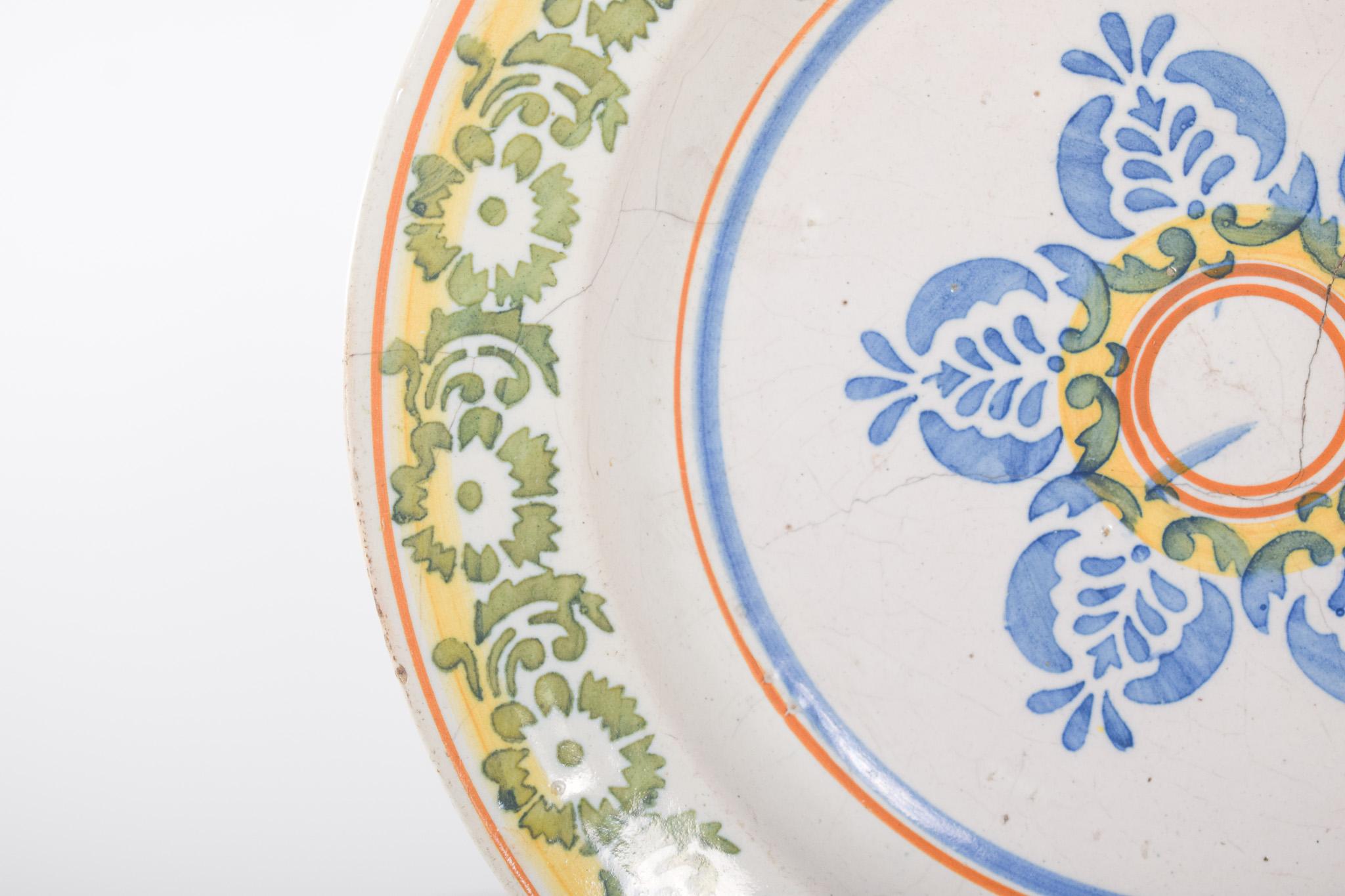 Plat creux en faïence portugaise, attribué à Coimbra. Décor polychrome avec au centre un motif floral symétrique dans les tons bleu, jaune et deux petits cercles orange. Le bord est décoré de fleurs et de feuilles stylisées en vert sur fond crème,
