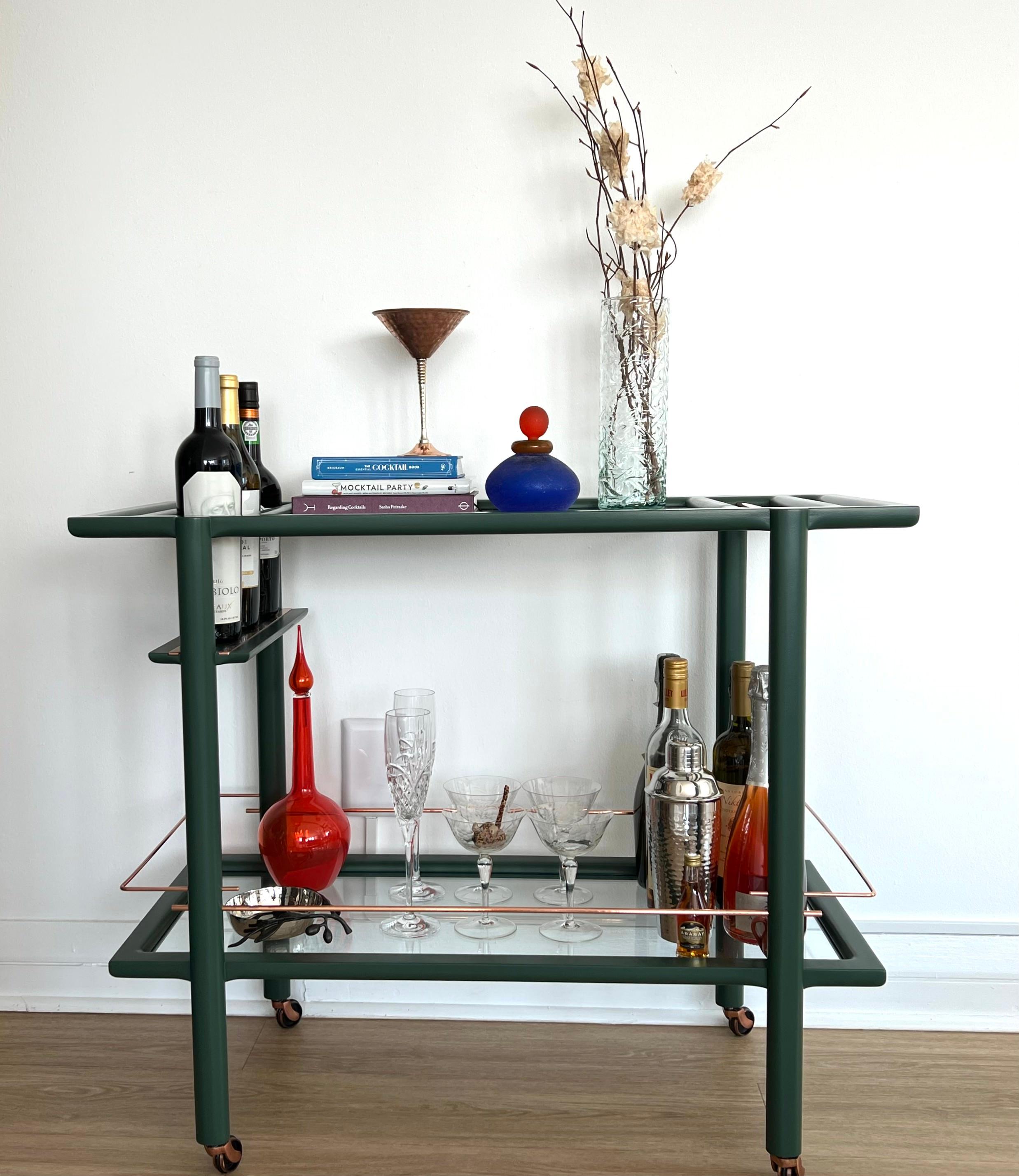 Ce chariot de bar est en stock et prêt à être expédié. Il est laqué sur mesure dans un vert profond, comporte des détails en cuivre et des étagères en verre fumé trempé. Ce chariot de bar en bois massif avec menuiserie découpée à la main est