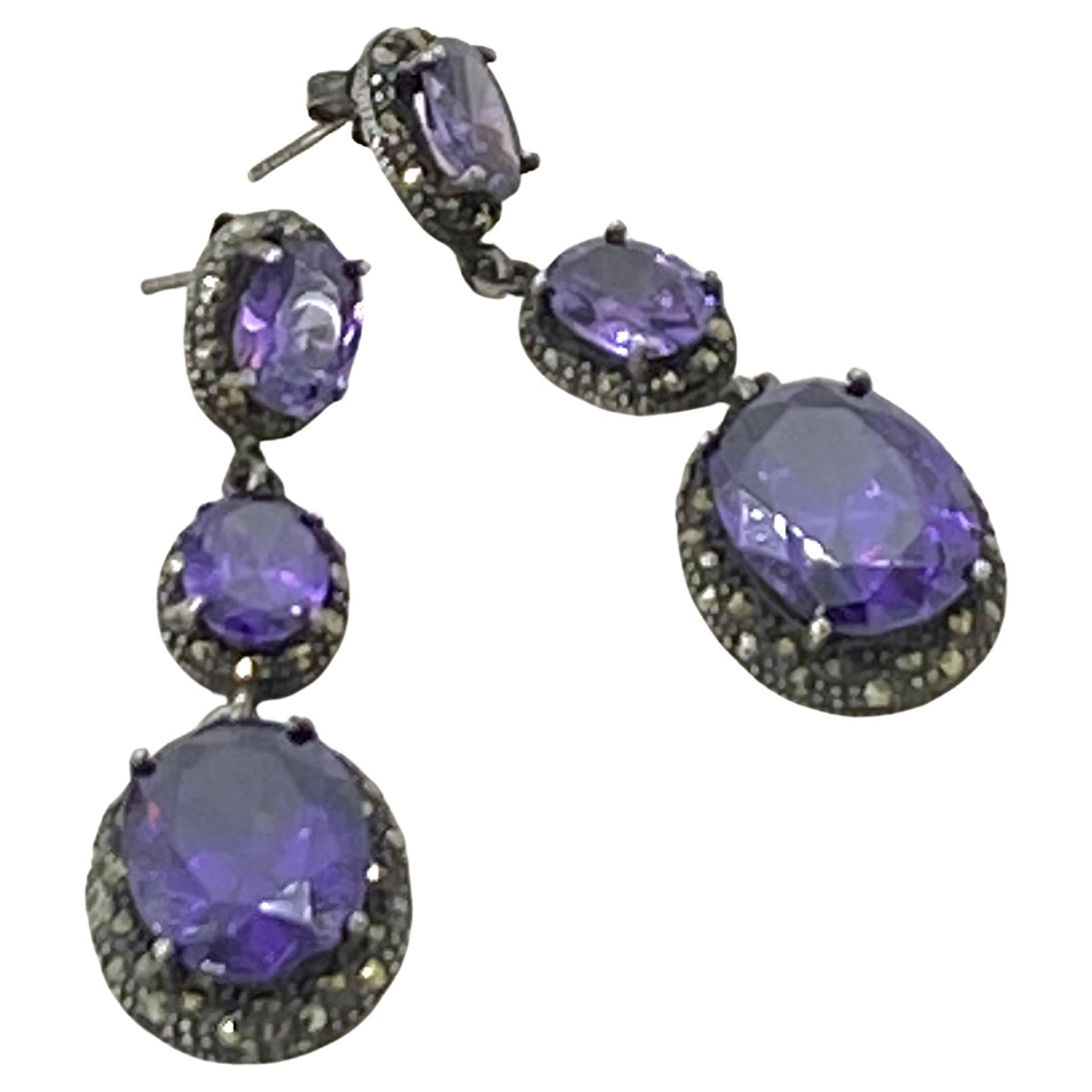 Si tratta di un paio di orecchini pendenti in cristallo e sterlina. Ci sono tre pietre di cristallo di iolite ovale di varie dimensioni collegate tra loro e incastonate in cornici di marcasite ossidata. Queste pietre viola intenso e gli orecchini in