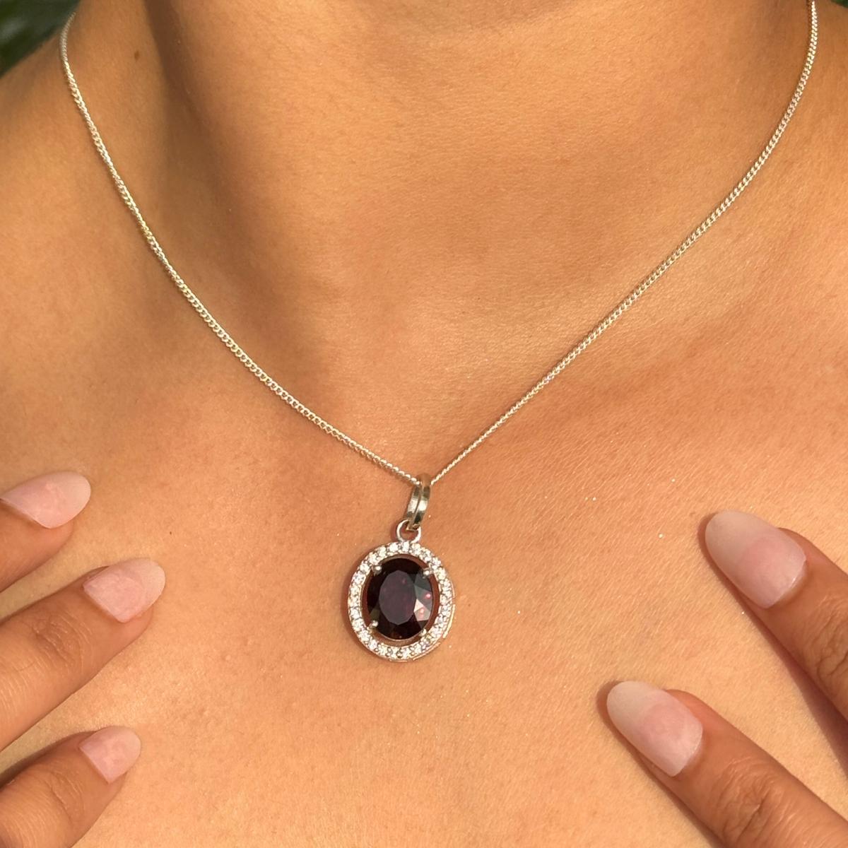 Ce pendentif en argent 925 est orné d'un grenat ovale d'un rouge profond. Entouré d'un délicat halo de zircon cubique scintillant, le pendentif est suspendu à une attache et à une chaîne en argent, rayonnant à la fois de raffinement et d'éclat.
