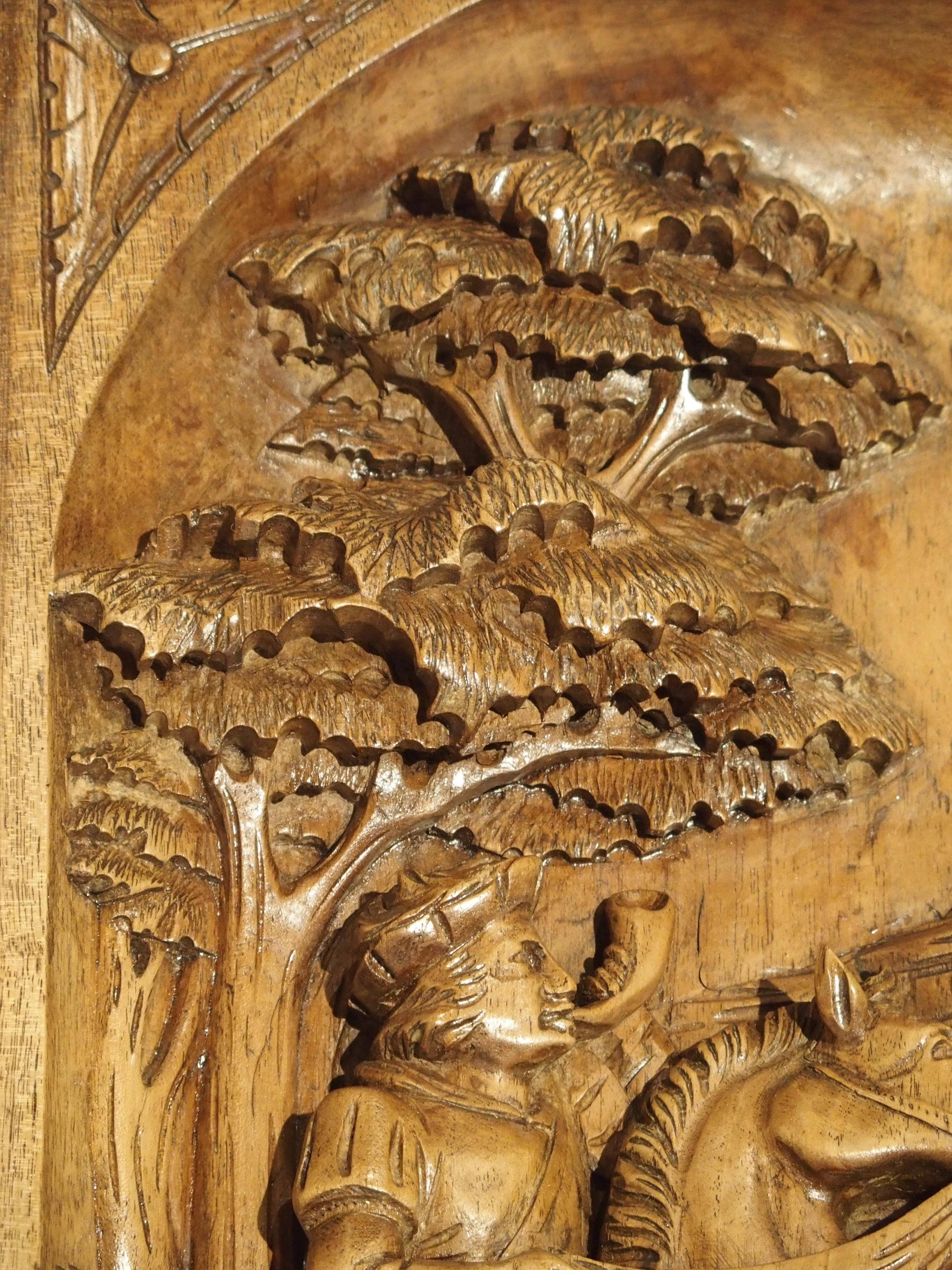 From France, this hand carved wooden plaque features deep and highly detailed carvings. It portrays a hunting scene with a man on a horse and a deer just below him. The man has a horn in his mouth, and his two hunting dogs are already engaged with