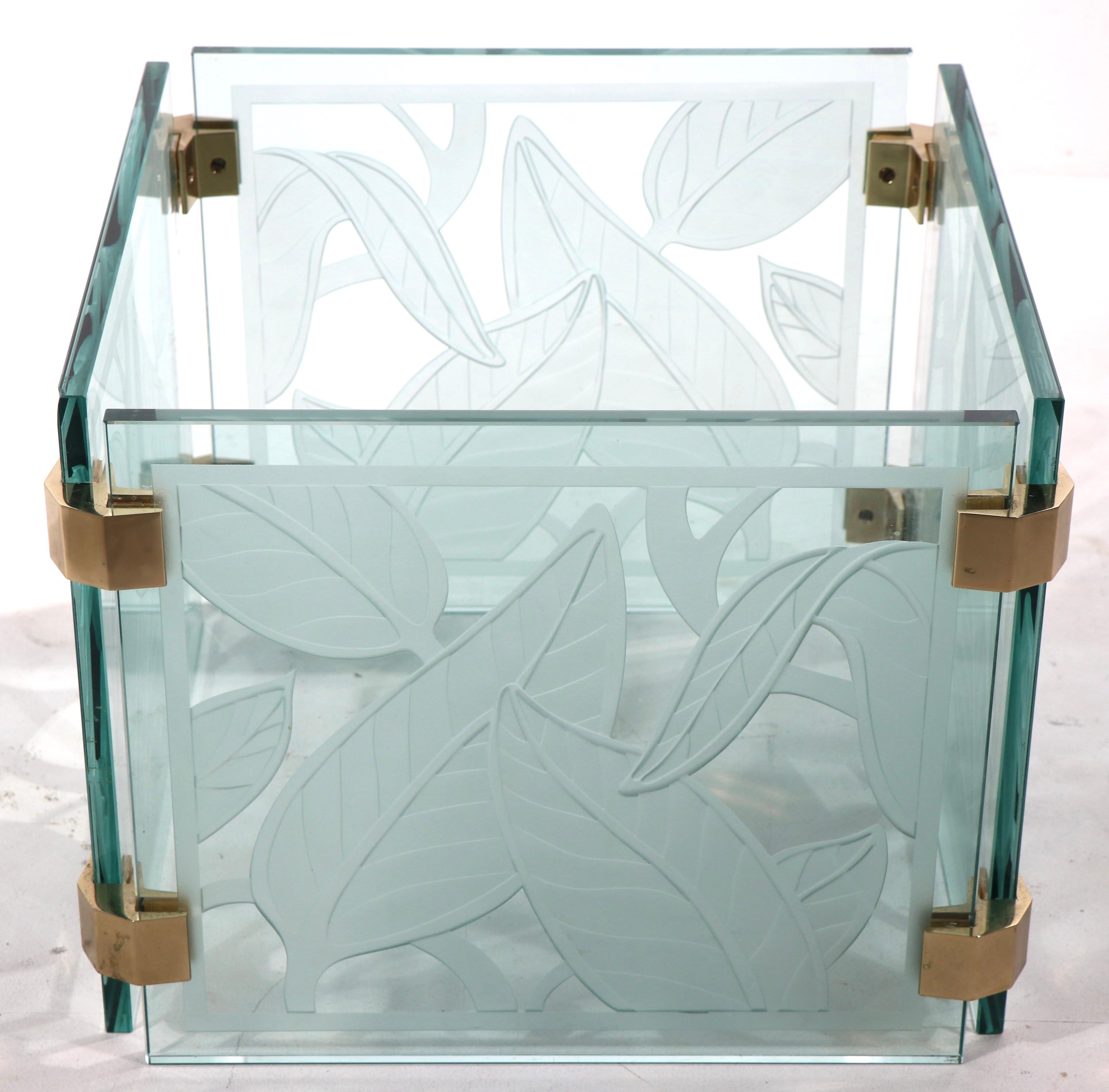 Chic voguish style modern glass coffee table having a thick ( 3/8 in ) rectangular beveled top which rests on square segmented base. The base consists of four square panels of deeply etched tropical foliate design, joined with brass fittings.
 Base