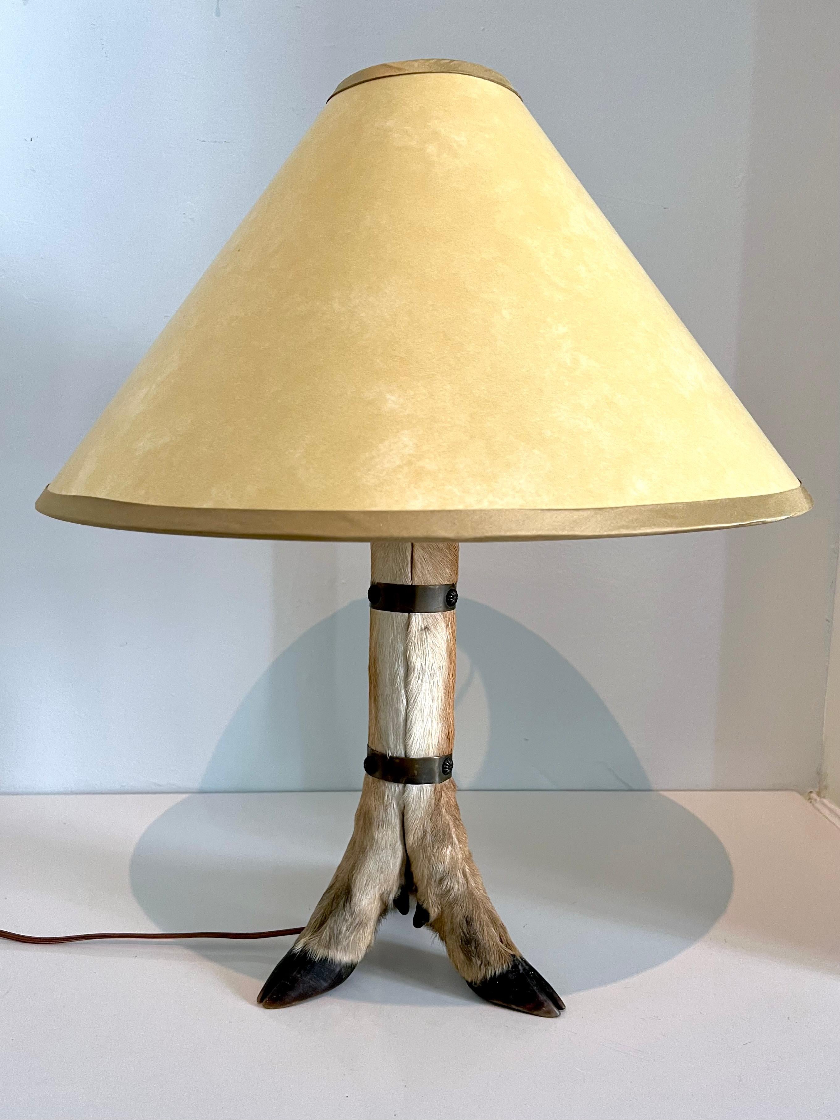 Unique table lamp featuring three taxidermy deer legs joined by blackened brass straps. This piece has been outfitted with a new custom parchment lampshade, In the style of Ralph Lauren - ready for your cabin or sports room. 

Measures: Base: 
8