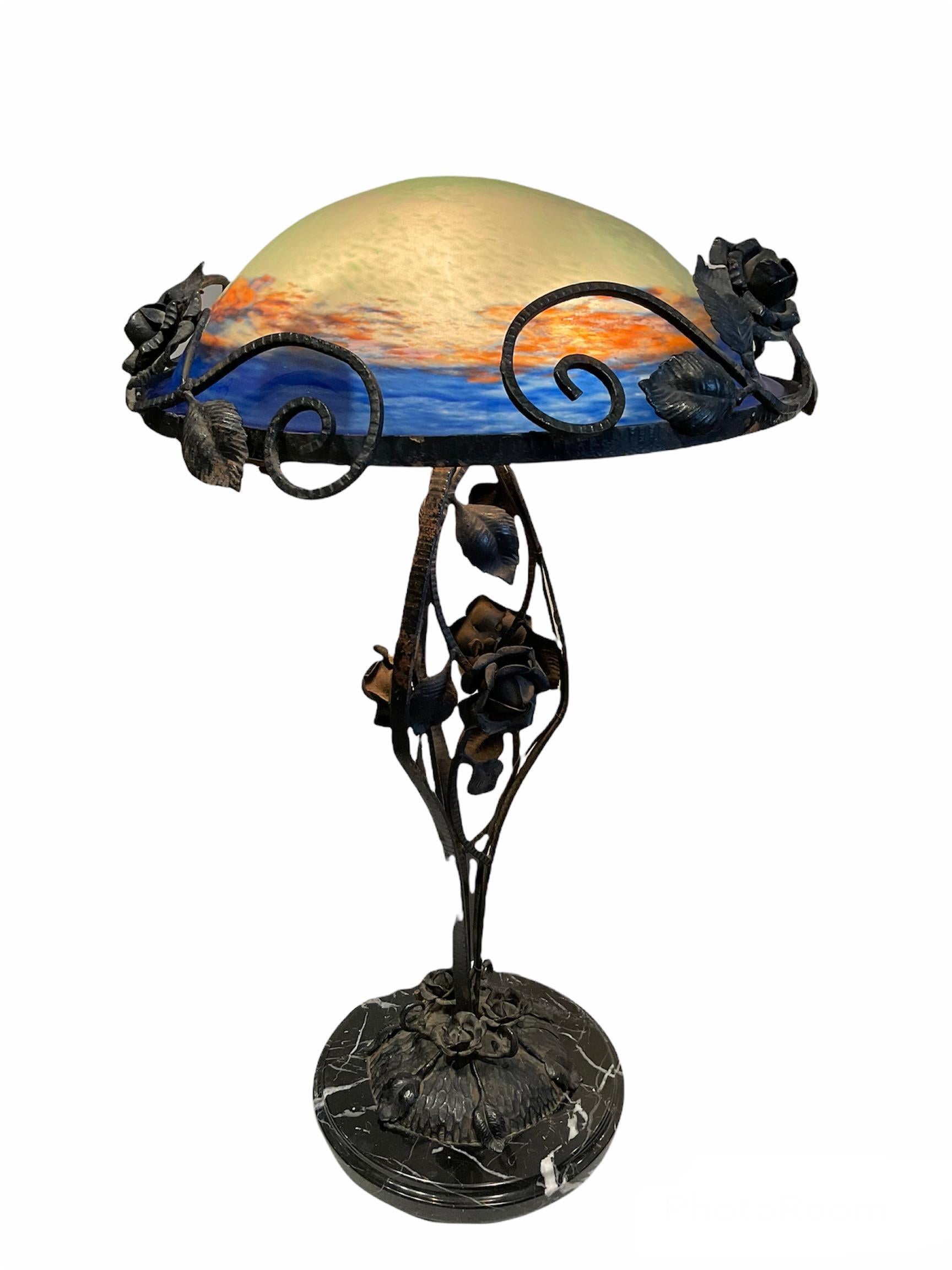 This is a hand painted half moon shaped glass paste shade lamp mounted in a wrought iron stem branches of roses and leaves. The half moon shade depicts an orange and royal blue colors brushstrokes scene of what appears to be a sunset. It stands over