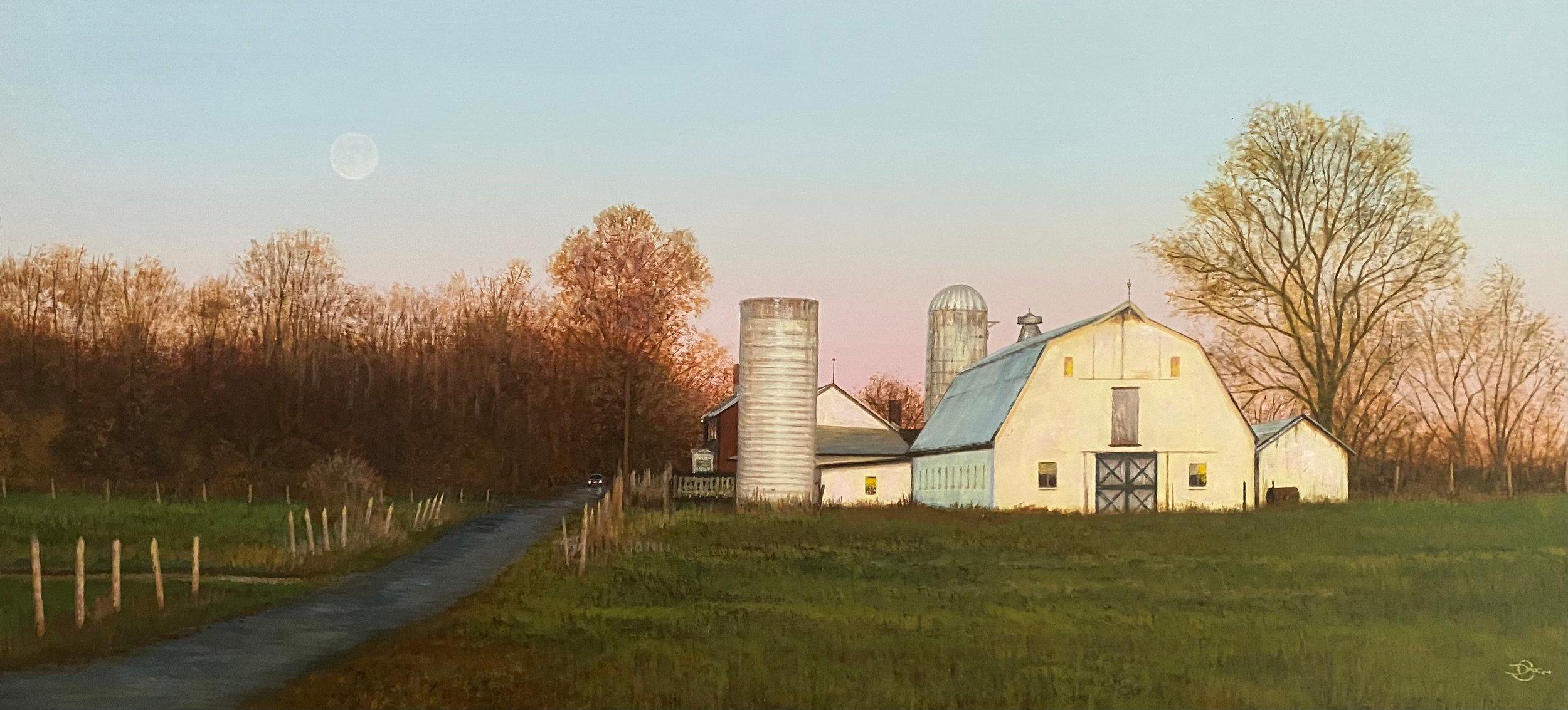 This piece, "Coming Evening", is a 10x21 acrylic painting on board by artist Del Bourree Bach featuring a rural landscape at dusk. Headlights glow down an old dirt road lined with pastures on both sides. A large white barn and silo soak in the last