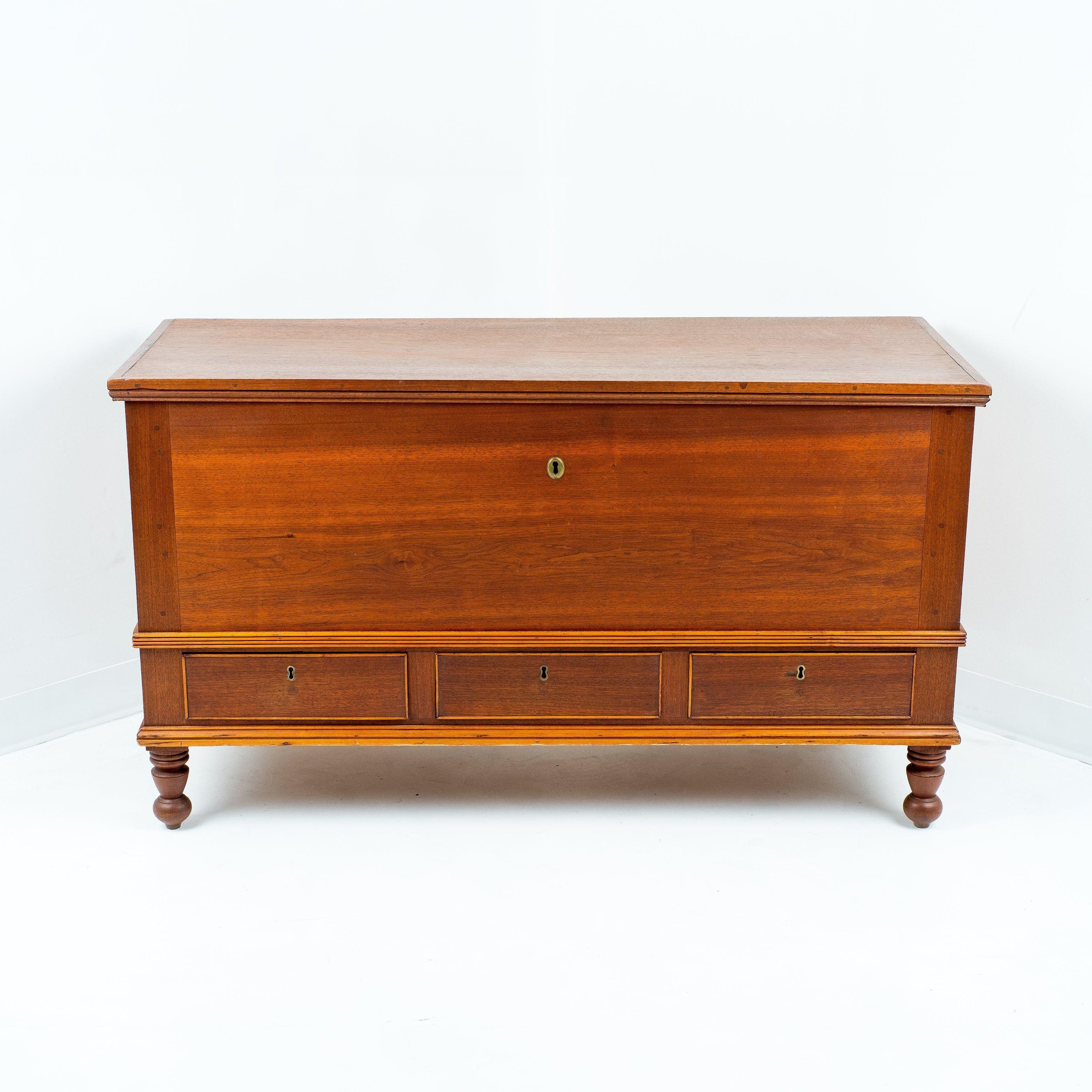 American black walnut dower chest over three divided drawers. The chest features post and panel construction with the corner posts terminating in turned feet. The hinged lift top reveals an interior fitted with till, and the top has an applied rim