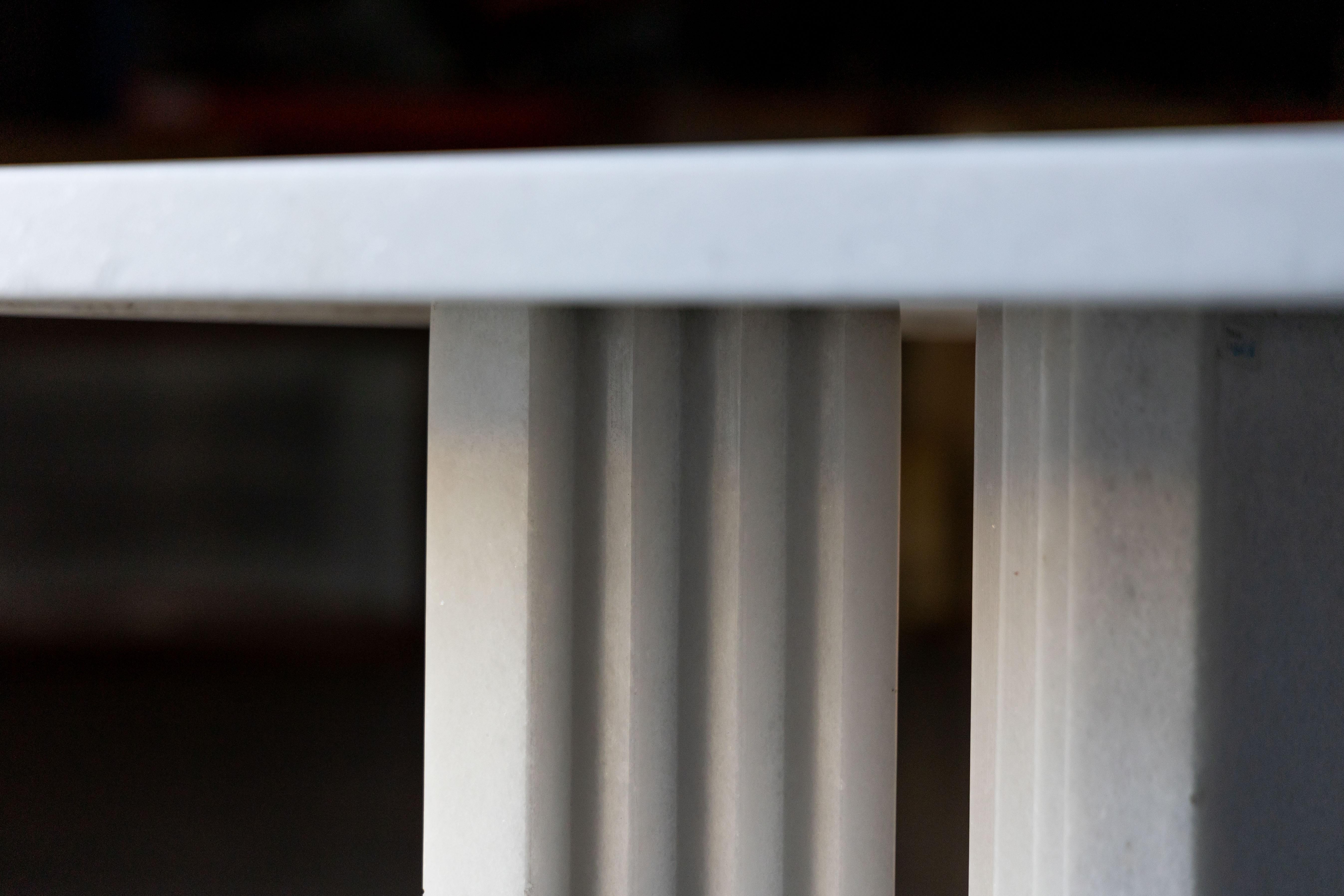 The 'Delfi' dining table, crafted entirely from pristine white marble, showcases its natural veining and impeccable beauty. The table features a striking rectangular top supported by two sculptural base elements, inspired by the contours of ancient