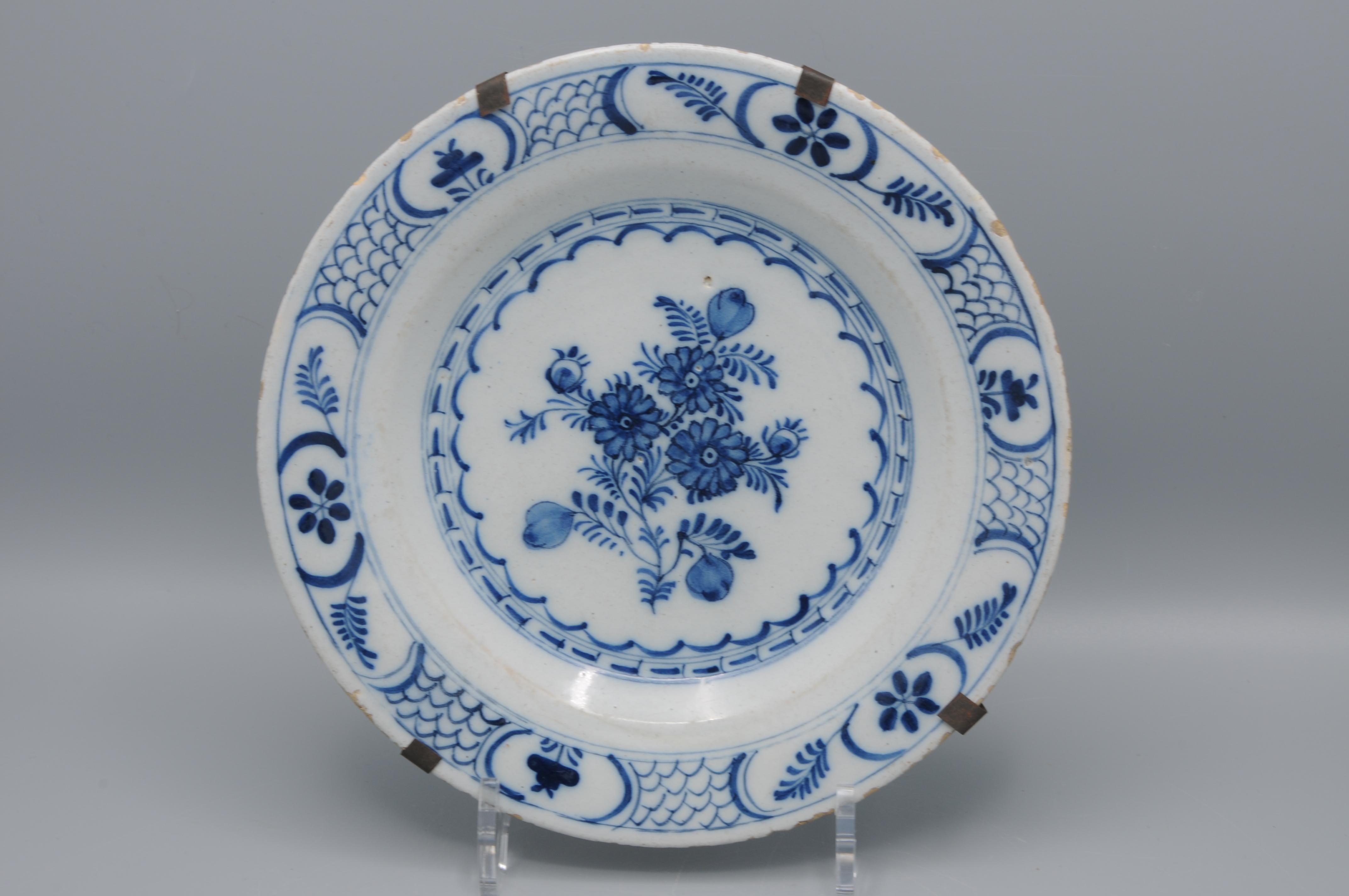 Assiette en faïence étamée de Delft décorée en bleu sur fond blanc, présentant un bouquet central de fleurs stylisées enfermées dans un médaillon festonné. Le cavetto et la bordure sont divisés en panneaux alternant des motifs floraux, des motifs en