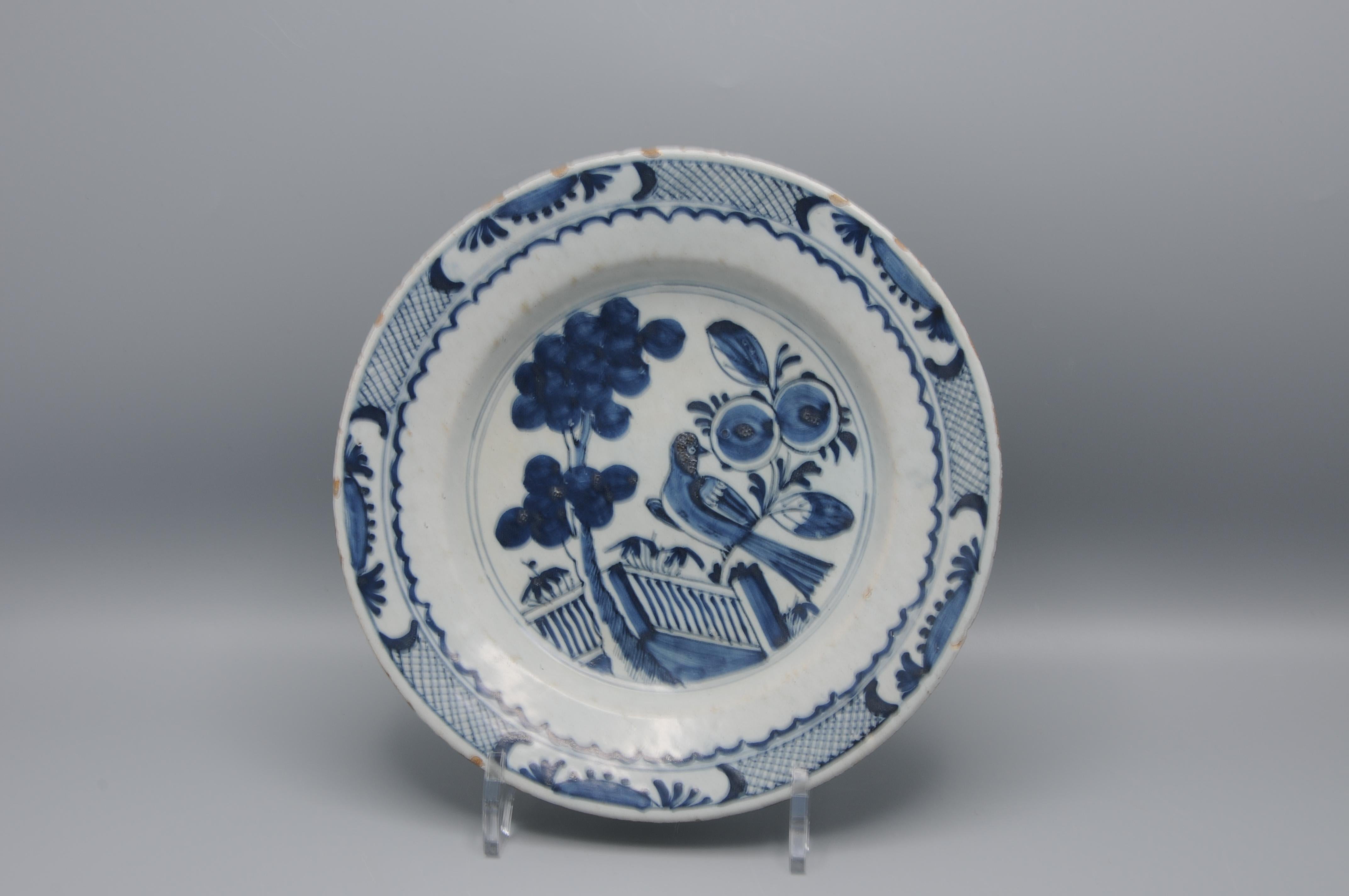 Assiette hollandaise en faïence étamée du milieu du 18e siècle, non marquée, décorée en bleu sous glaçure d'un motif de Chinoiserie. La scène centrale représente un oiseau perché sur une clôture, entouré de plantes à fleurs, d'arbustes et