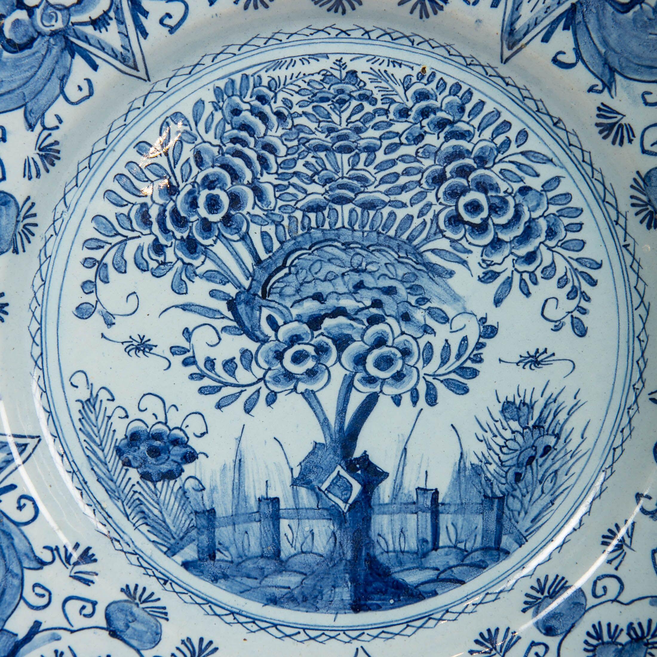 A Dutch Delft blue and white charger hand-painted showing a flowering tree in a fenced garden. Grasses, a single large flower, and insects are shown behind the fence. This design is contained within a circle formed by a chain of small crossed X's.