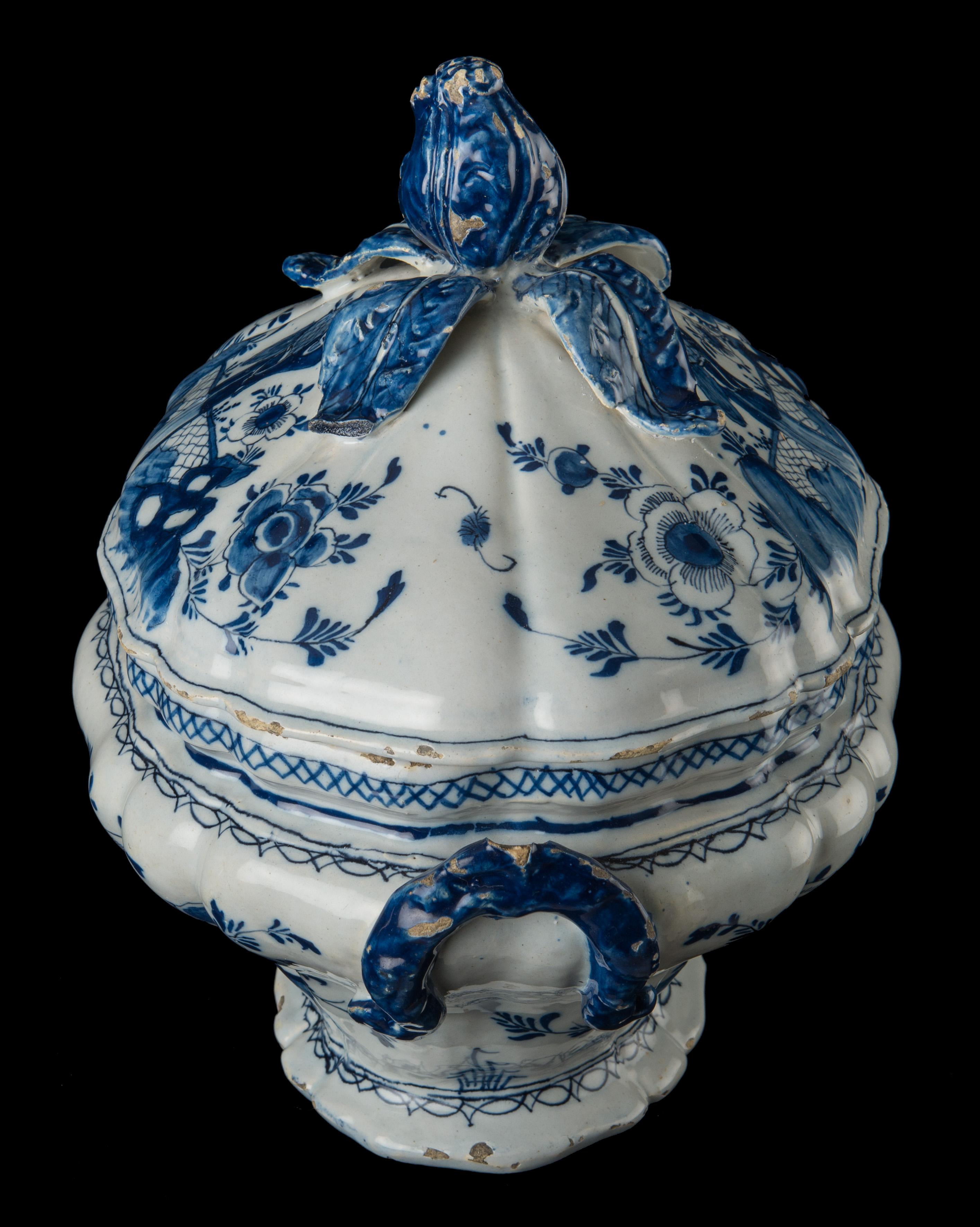 The blue and white oval-shaped tureen has a curved body on a spreading foot and two blue-colored foliate loop handles. The knob on the cover is modelled as a flower with four leaves. The tureen and cover are painted on either side with a Chinese