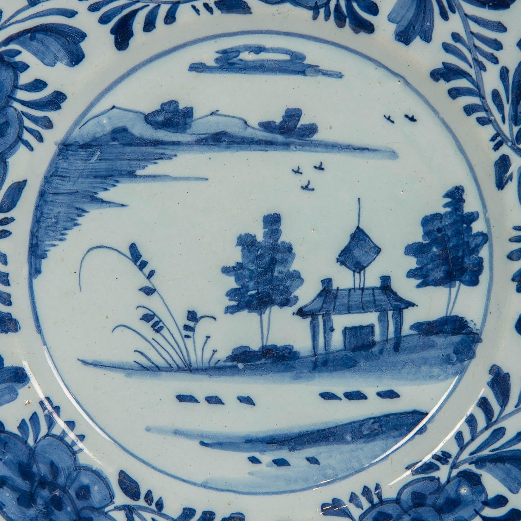 A Dutch Delft blue and white dish showing a waterside scene.
What makes this dish so lovely is the naively hand-painted scene showing a pair of stylized trees flanking a small house by the waterside.
Completing the decoration, the border is painted