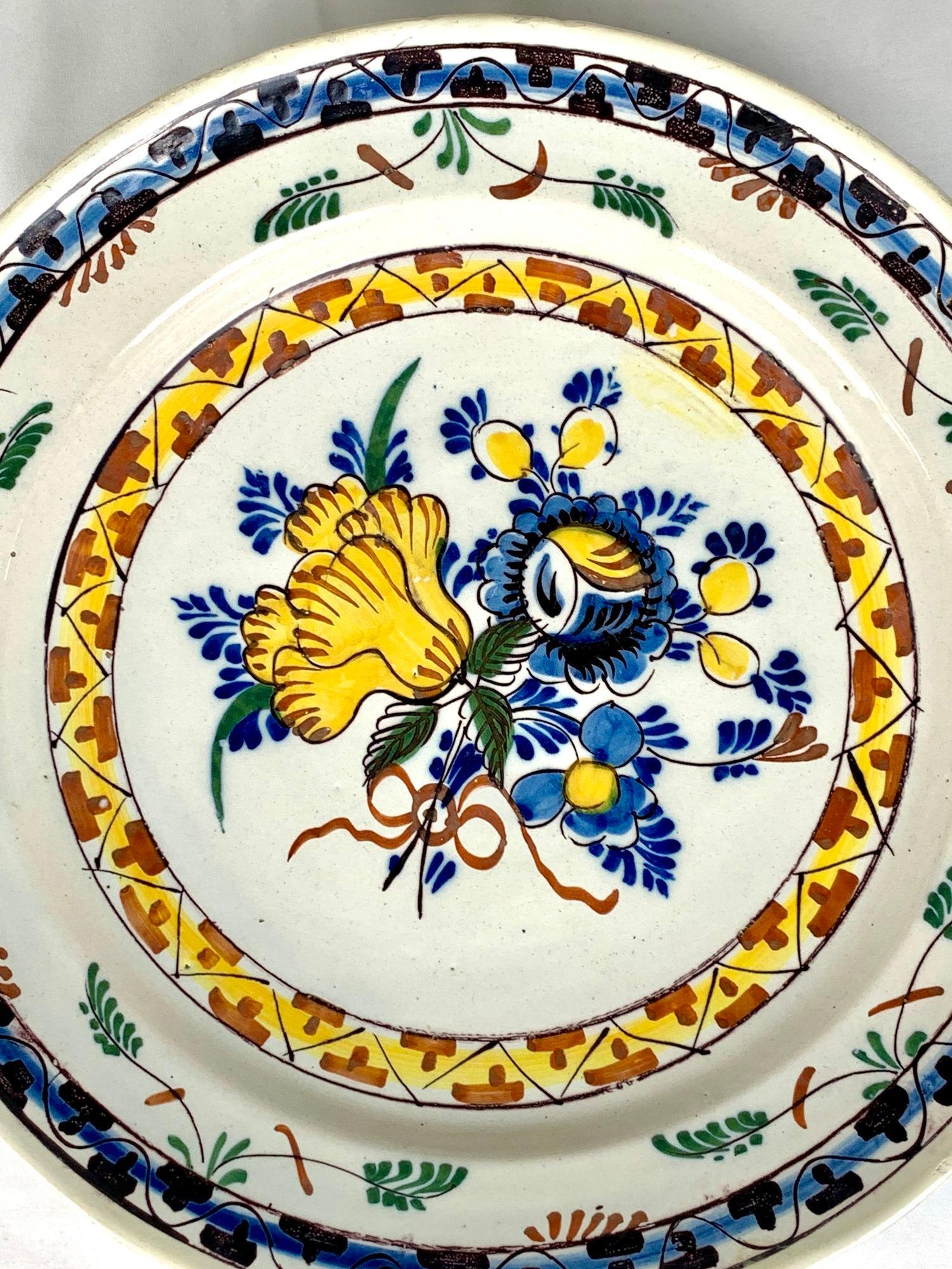 Made in the Netherlands circa 1780, this lovely Delft charger features a hand painted bouquet of beautiful flowers.
We see a large yellow tulip, yellow tulip buds, and bright blue leaves, all tied together with an iron red bow.
Encircling the