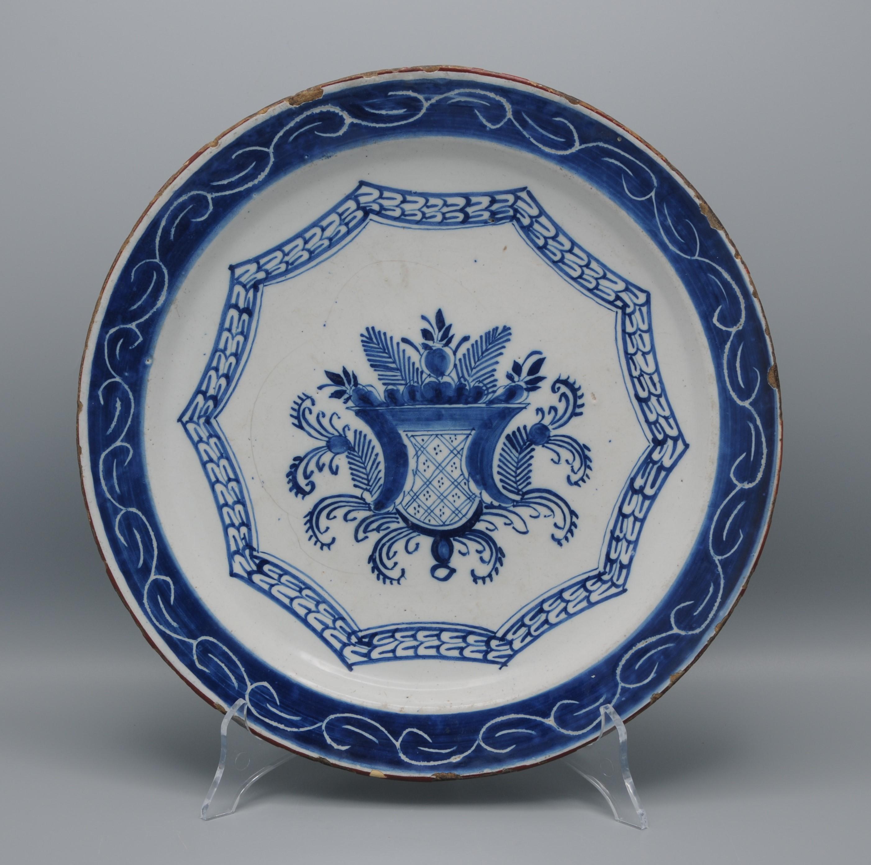 Piatto in ceramica blu della metà del XVIII secolo con decorazione in stile Daniel Marot di uno scudo tra fiori e volute di fogliame all'interno di un decagono. 
Bellissima decorazione del bordo in un ricco blu con volute in 'spare-technique'.
Non