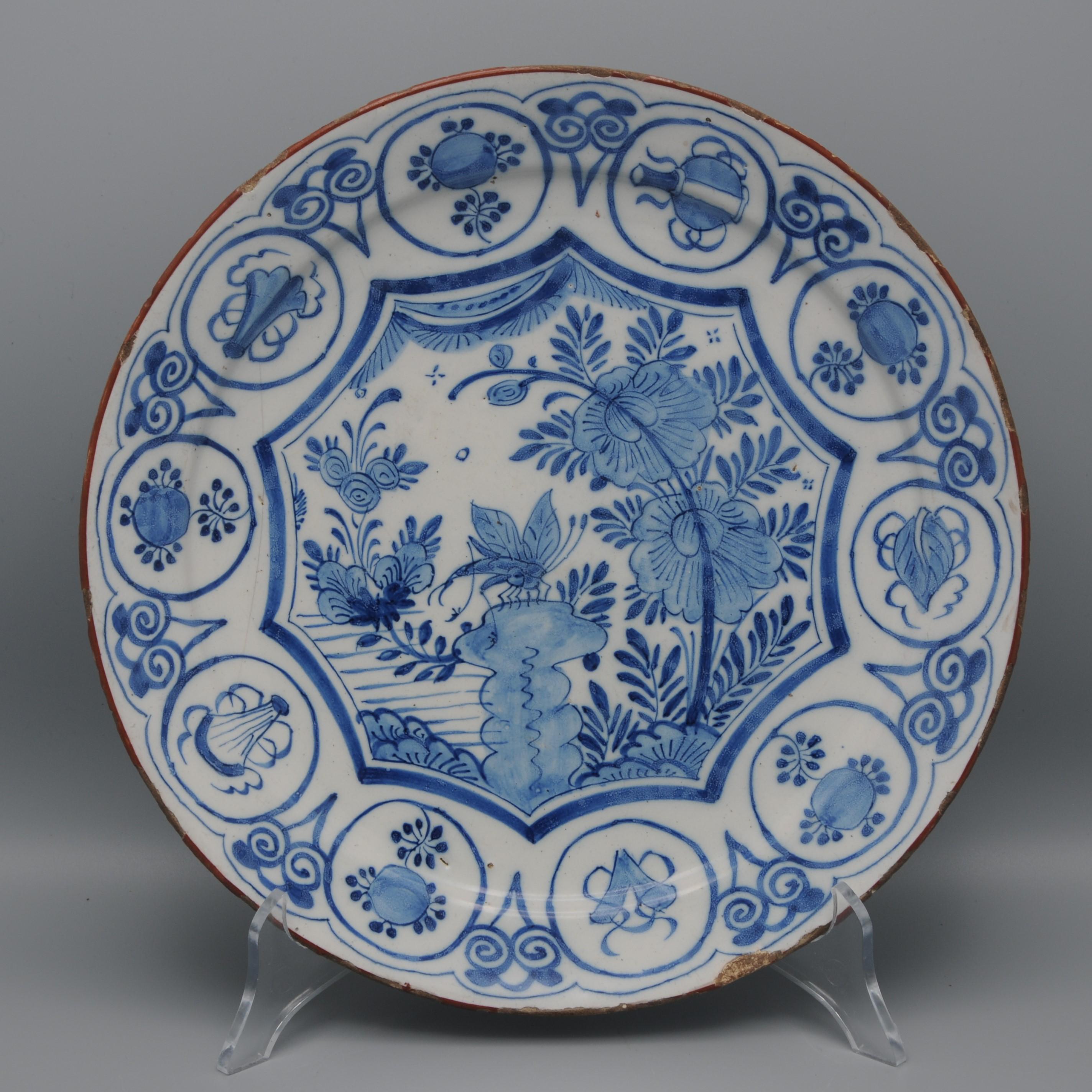 Chargeur de Chinoiserie de Delft représentant une grande libellule, dans un jardin chinois, assise sur un rocher. L'ensemble est entouré d'une bordure octogonale. Le bord est brun et la large bordure est ornée d'une série de rondeaux représentant