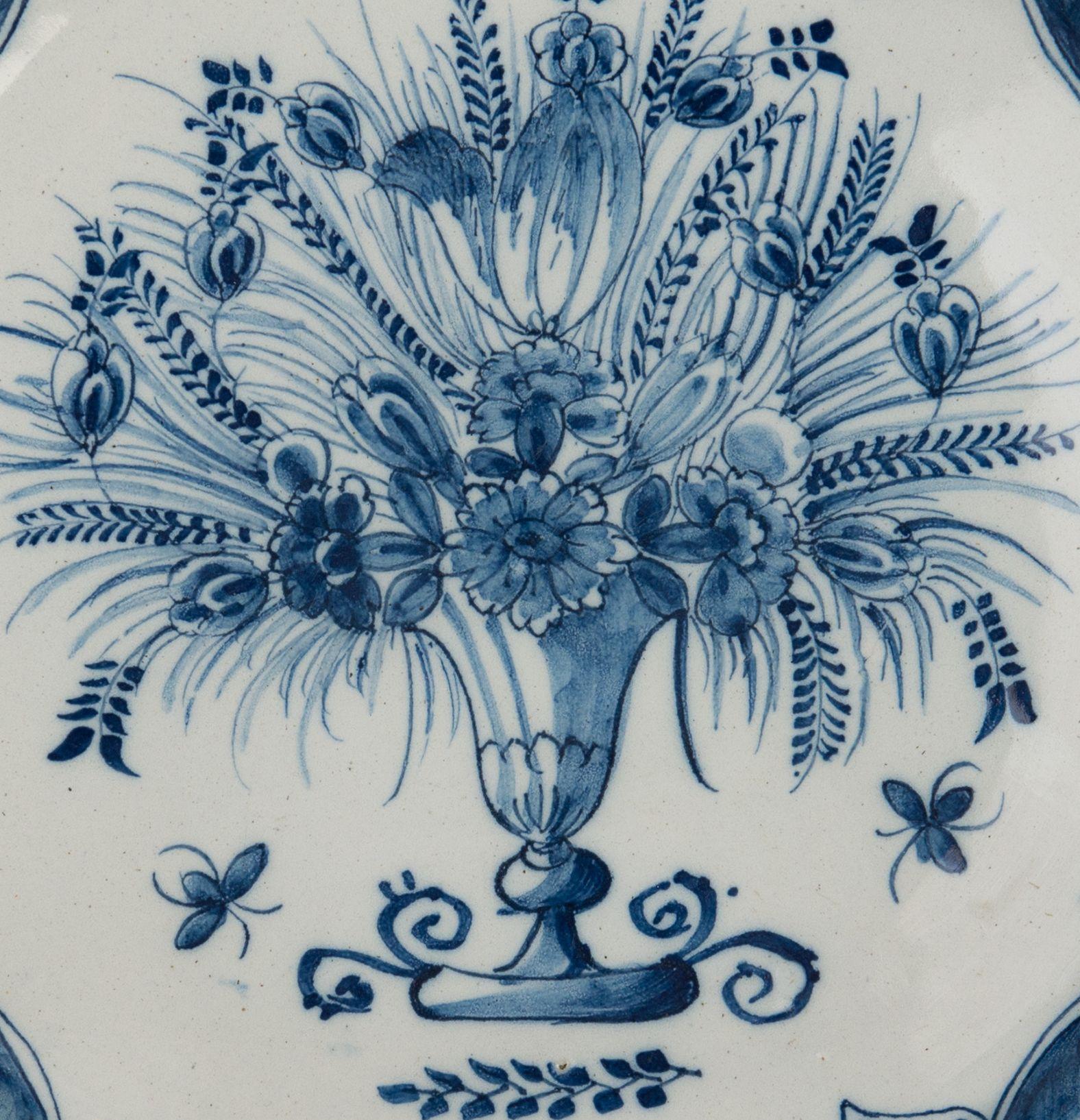 Blaue und weiße Schale mit Blumenvase. Delft, 1740-1760
Die Töpferei Three Bells. Zeichen: drei Glocken

Schale mit blau bemaltem Rand, der eine Vase mit mehreren Blumen und Zweigen zeigt. Unter der Vase ist ein kleiner Zweig abgebildet. Der Rand