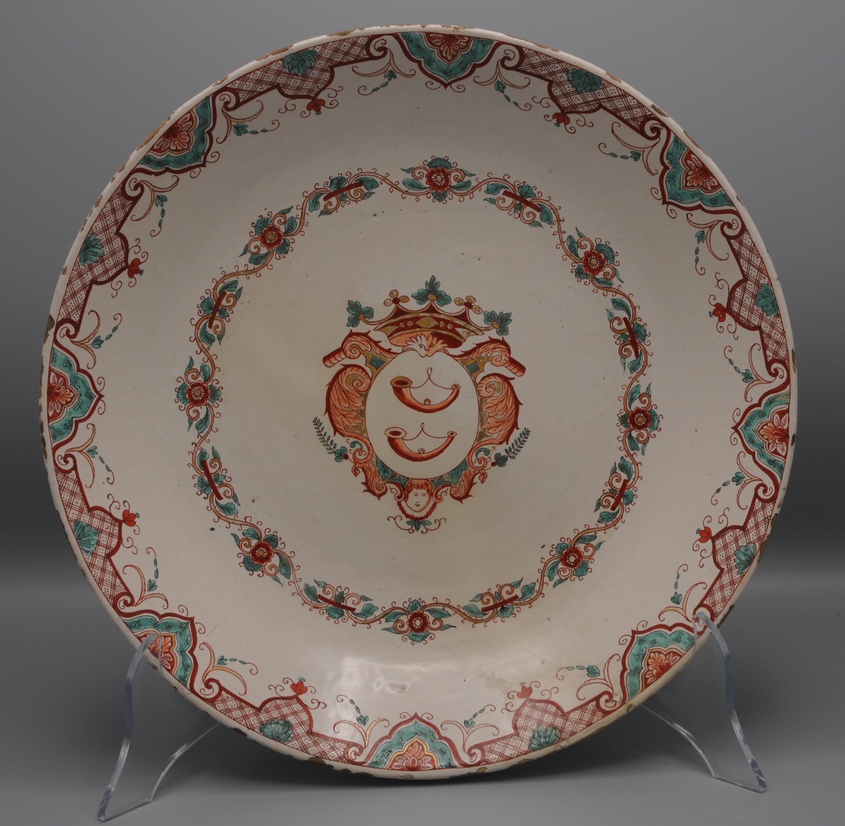 A Dutch Delft charger, decorated in petit-feu polychrome enamels and gilding, early 18th century. The central shield, painted with three hunting horns, is surmounted by a coronet and flanked by scrolling mantling with a cherub mask below. The arms
