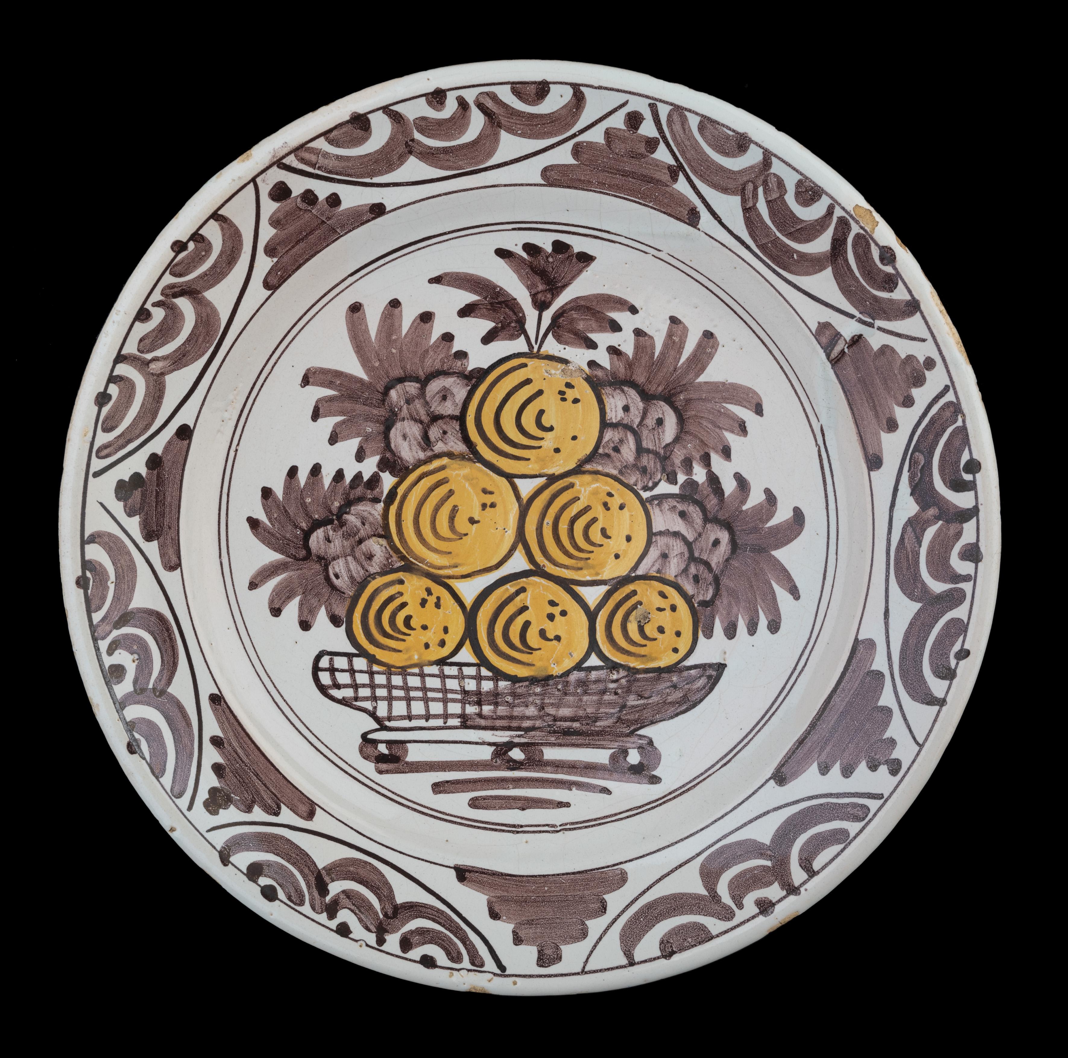 Dish with fruit in purple and yellow The Netherlands, 1660-1700.

The dish has a spreading, slightly raised flange and is painted in purple and yellow with a fruit decor of grapes and apples or oranges, set in a double circle. The well is