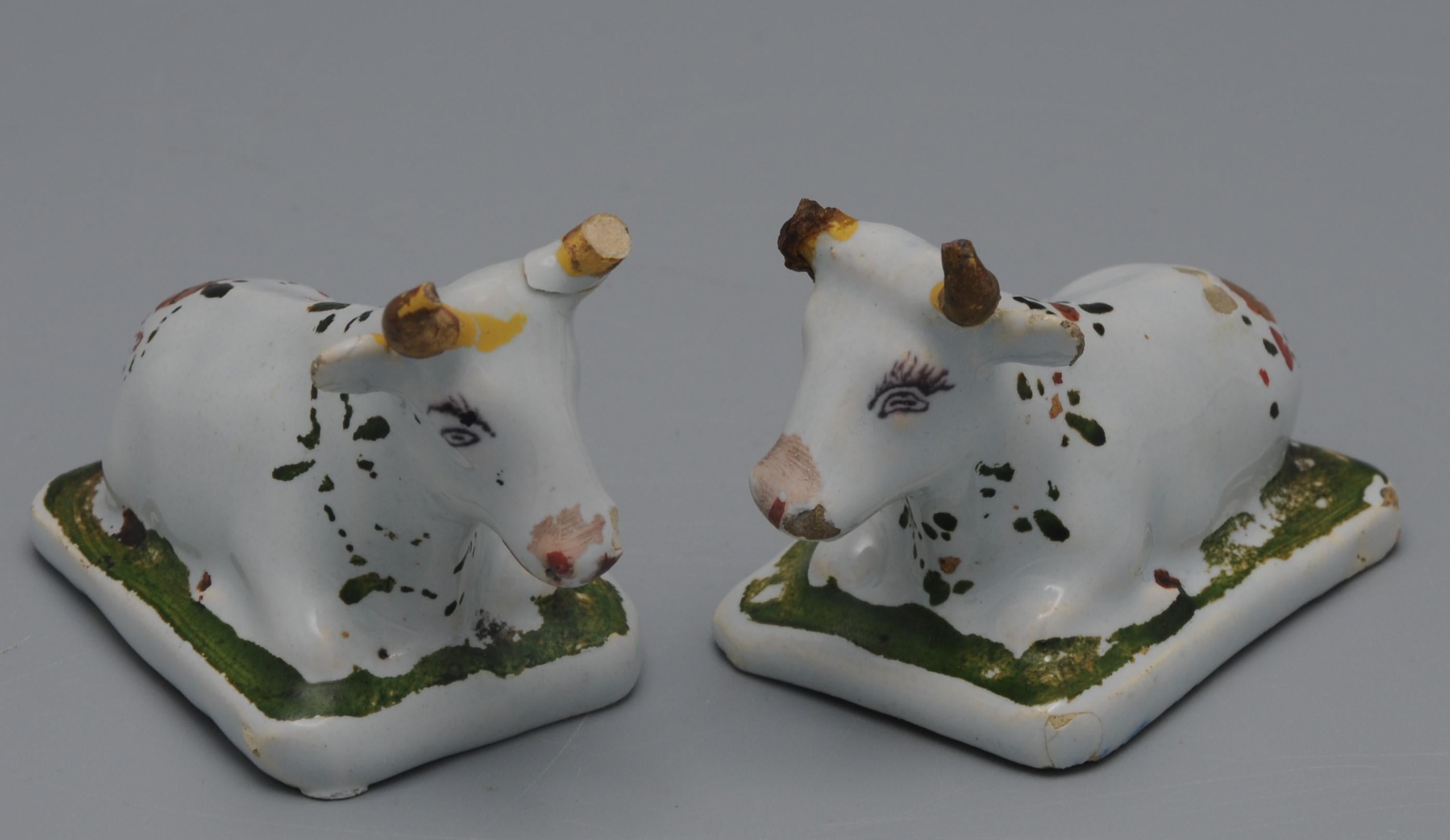Pair of minature white Delft recumbent cows, modelled affronté with their heads towards each other. Remains of cold painting (overglaze) of green on the plinth and floral wreaths and gilt horns. Adoring cows or bulls with floral wreaths harks back