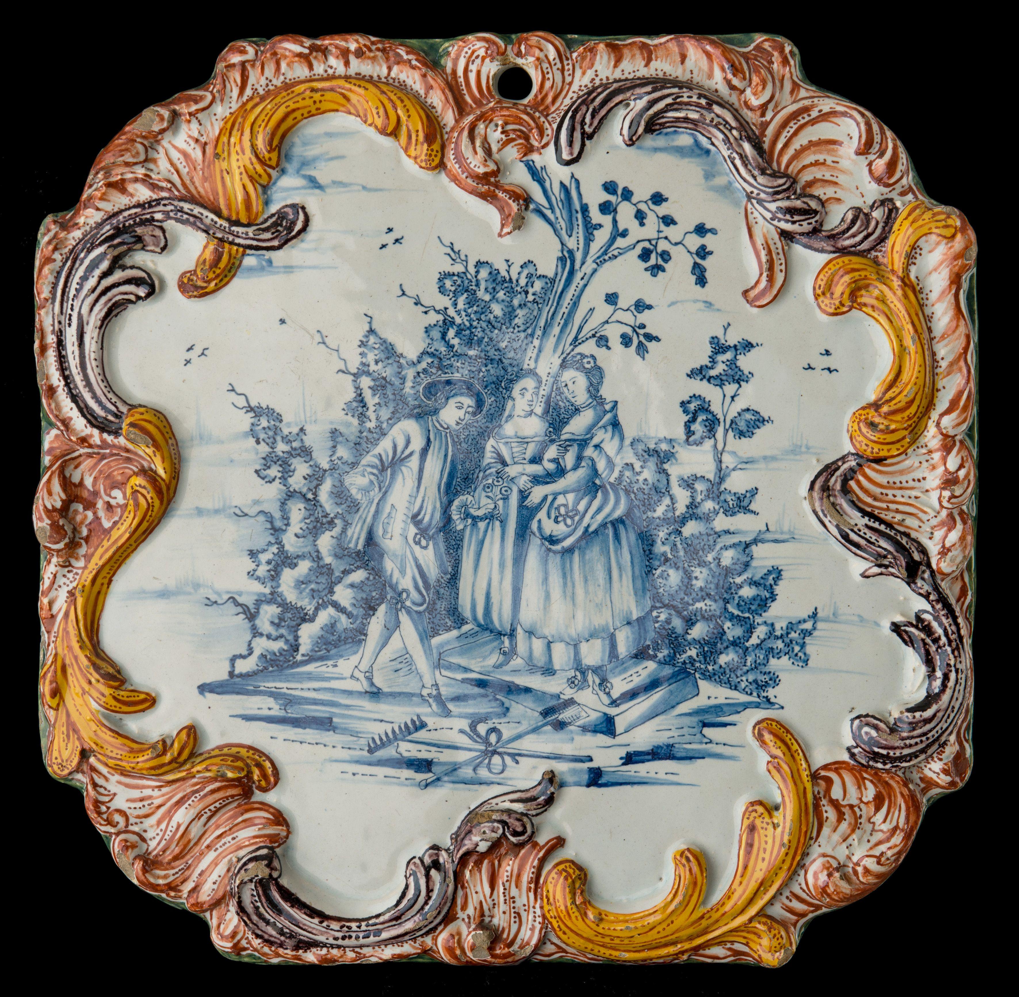 The octagonal plaque is painted in blue with a courteous scene. A young man offers a flower to a young lady accompanied by her chaperon, against a background of trees and shrubbery. On the foreground lie a rake and a shovel in cross shape, tied with