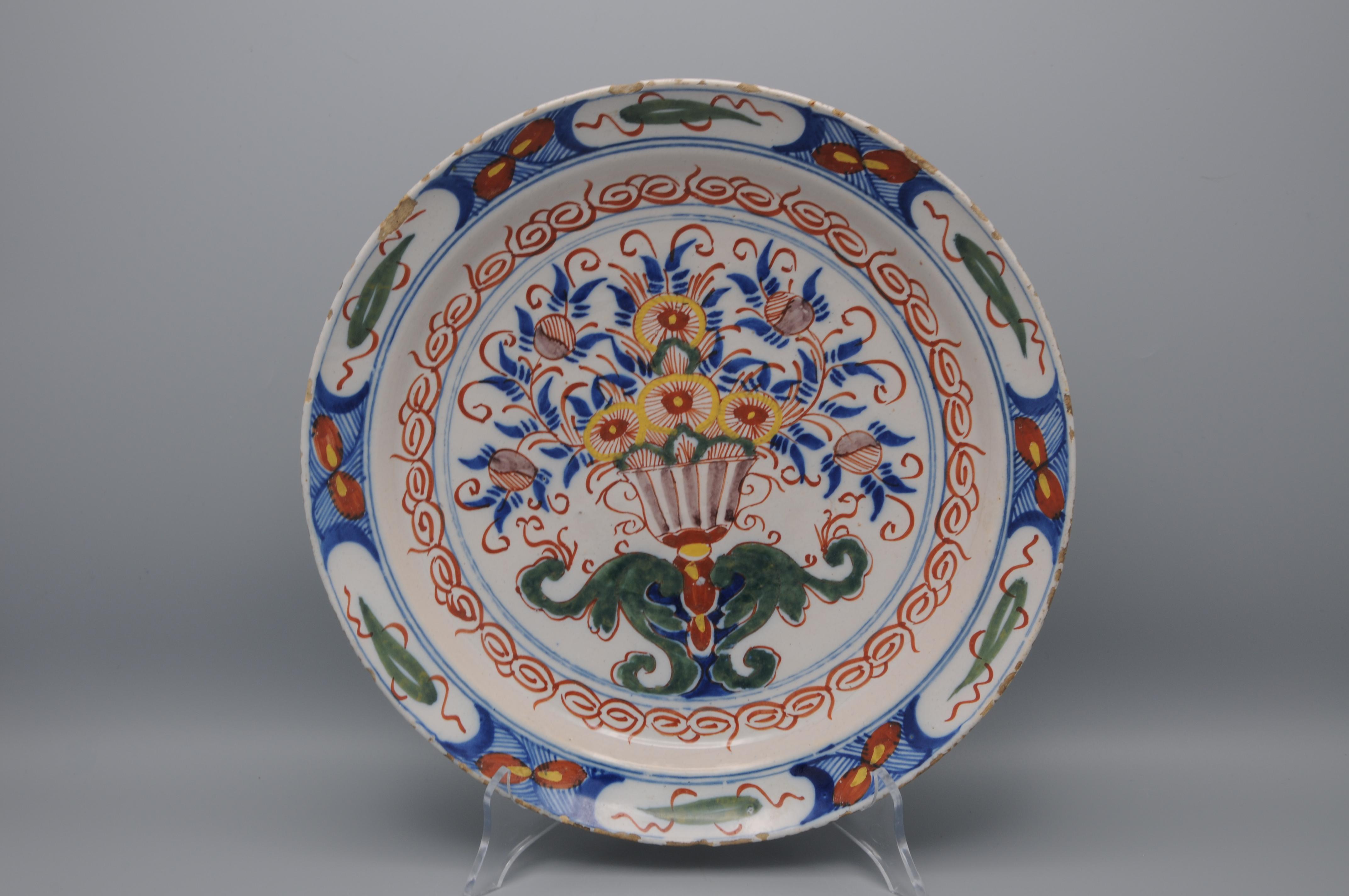 Plato polícromo holandés de Delft con animada decoración polícroma de jarrón central con abundancia de flores en la parte superior op volutas de follaje verde. 
Borde muy ornamentado con hojas estilizadas de Artemisa, derivado de los motivos chinos