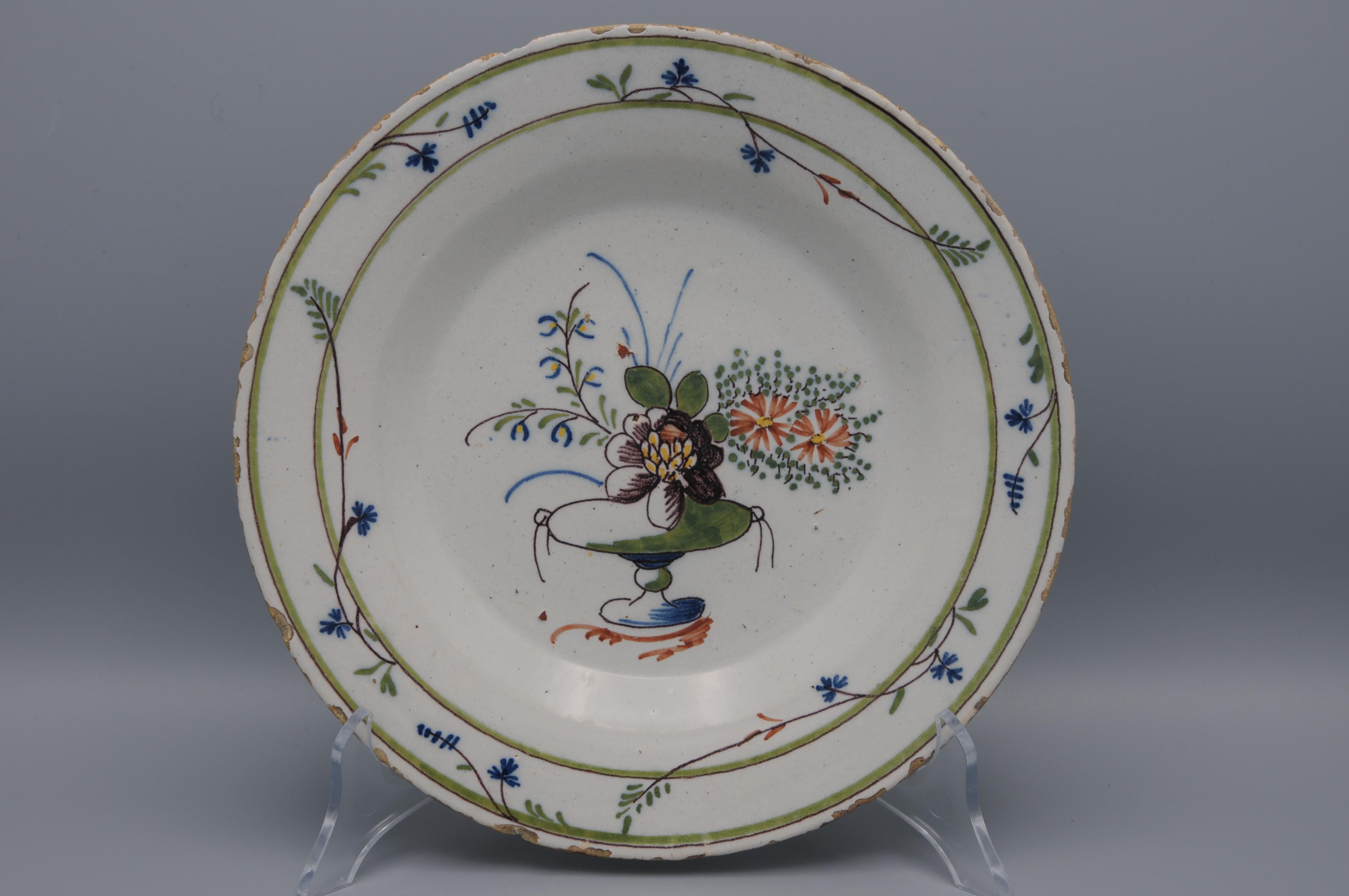 Late 18th century Delftware plate with Neoclassical decoration of a central vase with manganese flower and flowering branches. The border is decorated with intertwining cornflowers. 

Cornflowers were the favorite flower of Mary Antoinette. Sevres