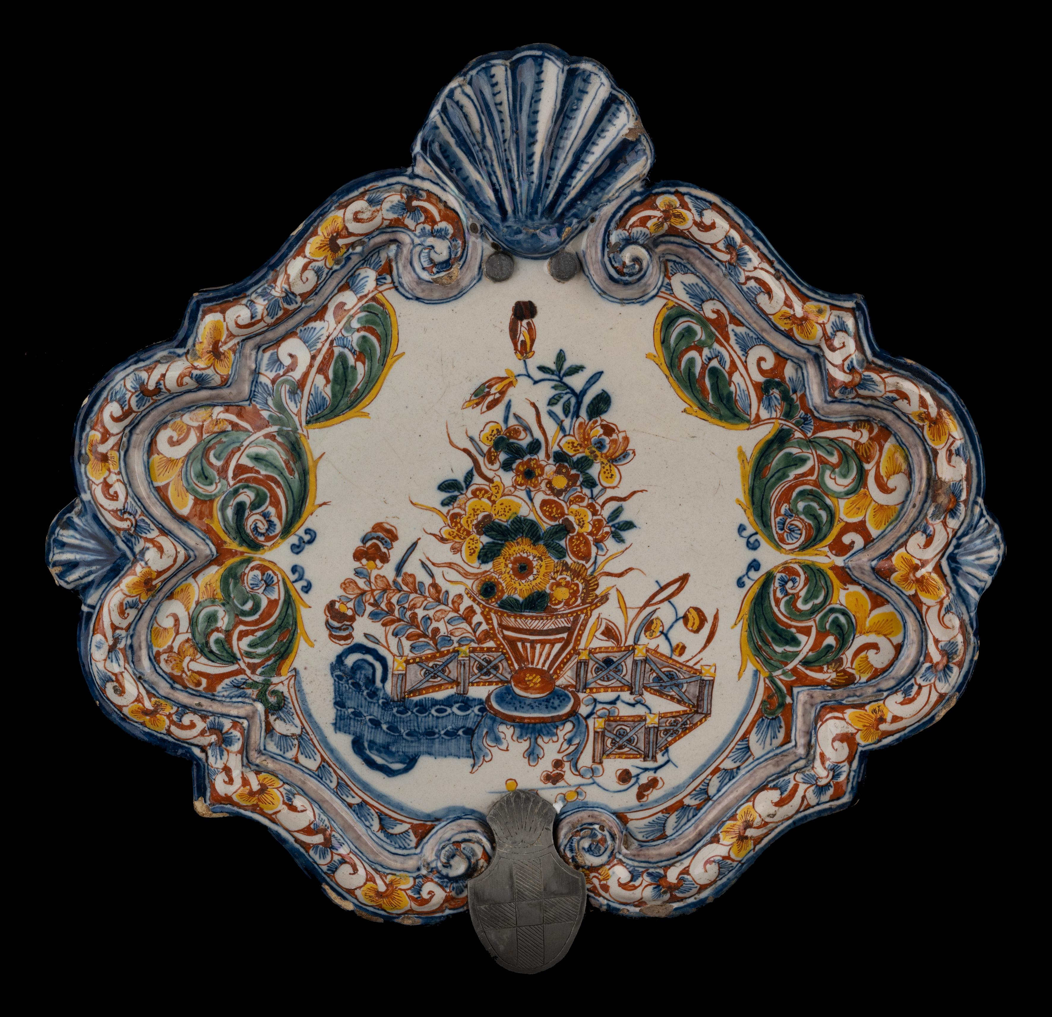 Polychrome plaque with a flower vase. Delft, 1740-1760

The lozenge-shaped plaque has a raised, accolade-shaped rim interrupted at the top by a large shell and at the bottom by a smaller shell. A shell is also modelled on both sides. The plaque is