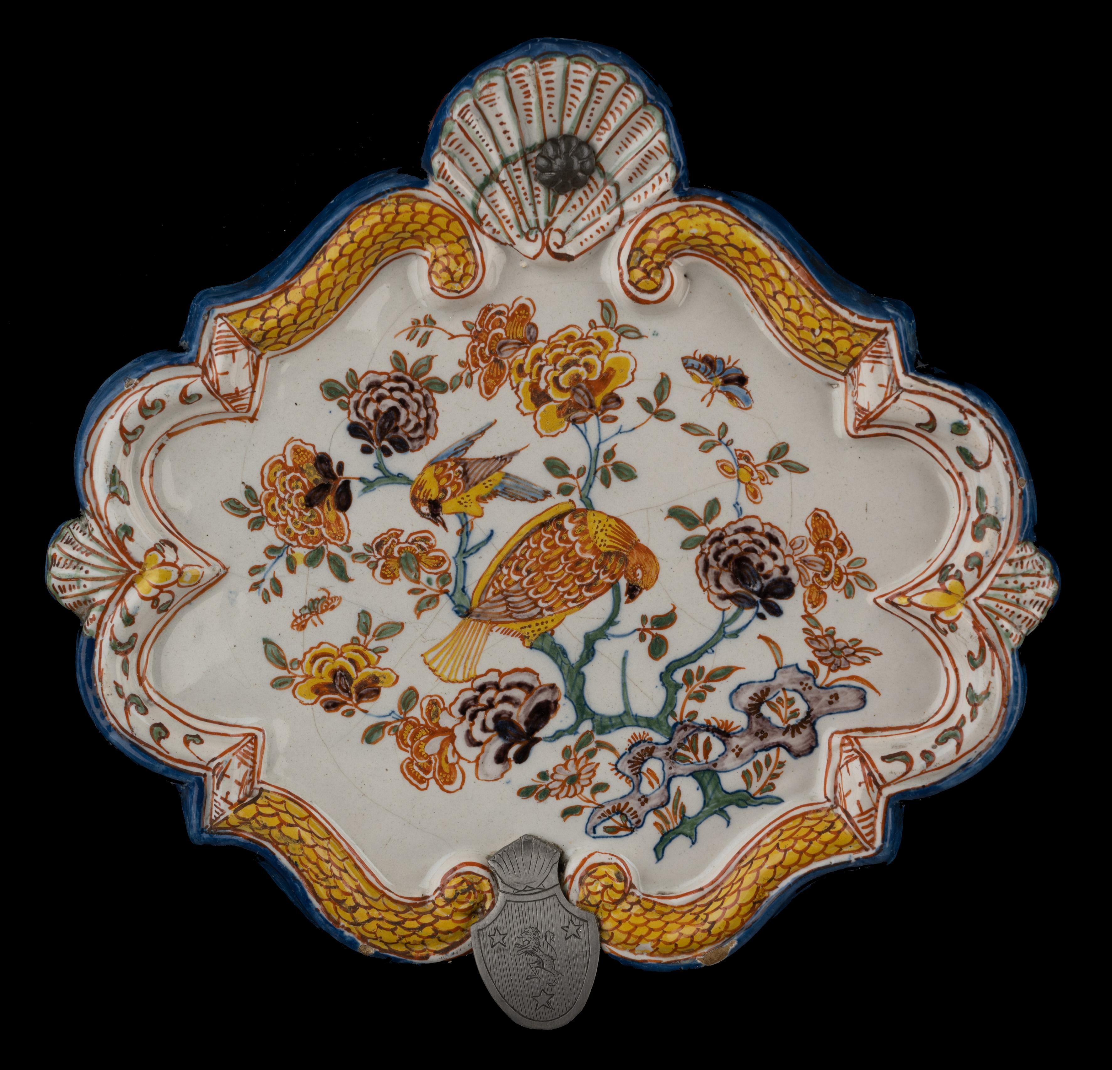 Polychrome plaque with floral chinoiserie decor. Delft, 1740-1760

The lozenge-shaped plaque has a raised, accolade-shaped rim interrupted at the top by a large shell with suspension hole and at the bottom by a smaller shell. A shell is also