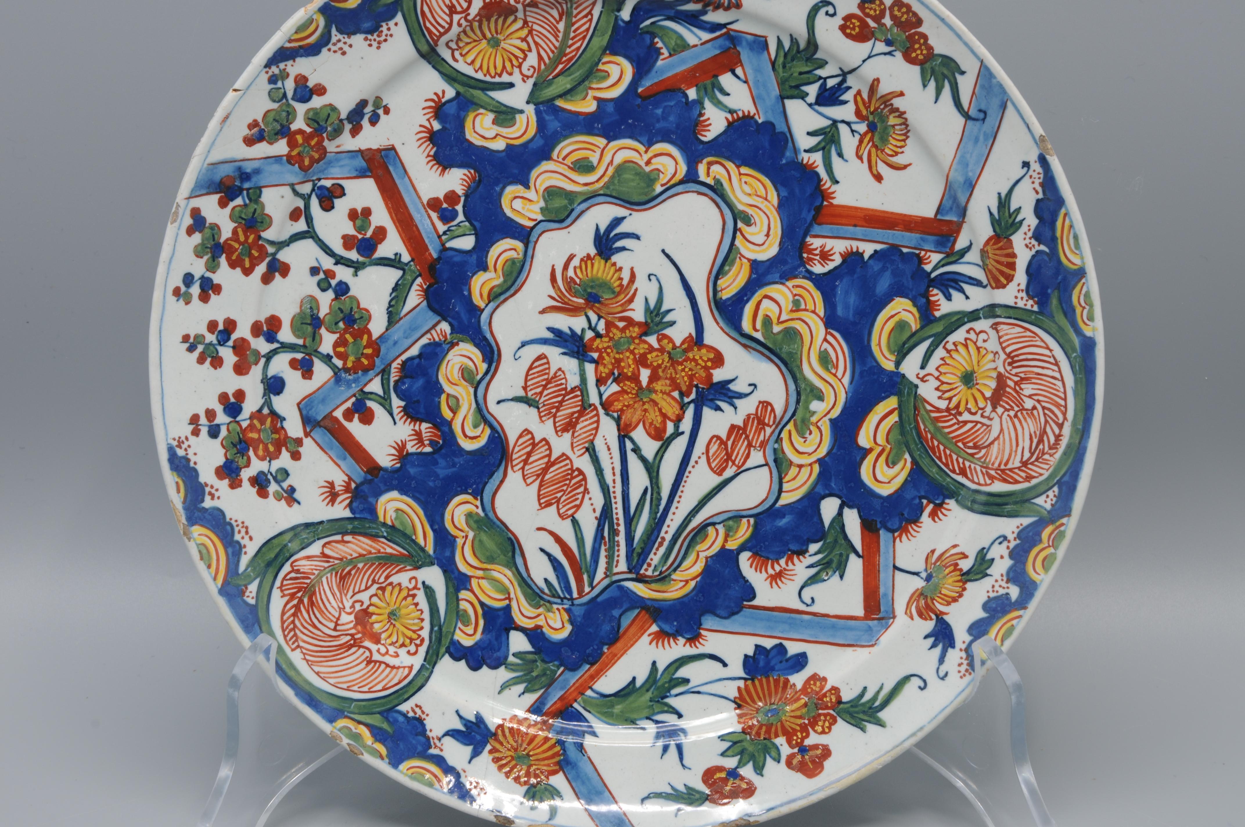 A polychrome Dutch Delft earthenware dish, traditionally called a “bliksem” (lightning) dish, painted in vibrant colors with a shaped central cartouche reserved with waterflowers, surrounded by characteristic zigzag motifs and monograms. The design