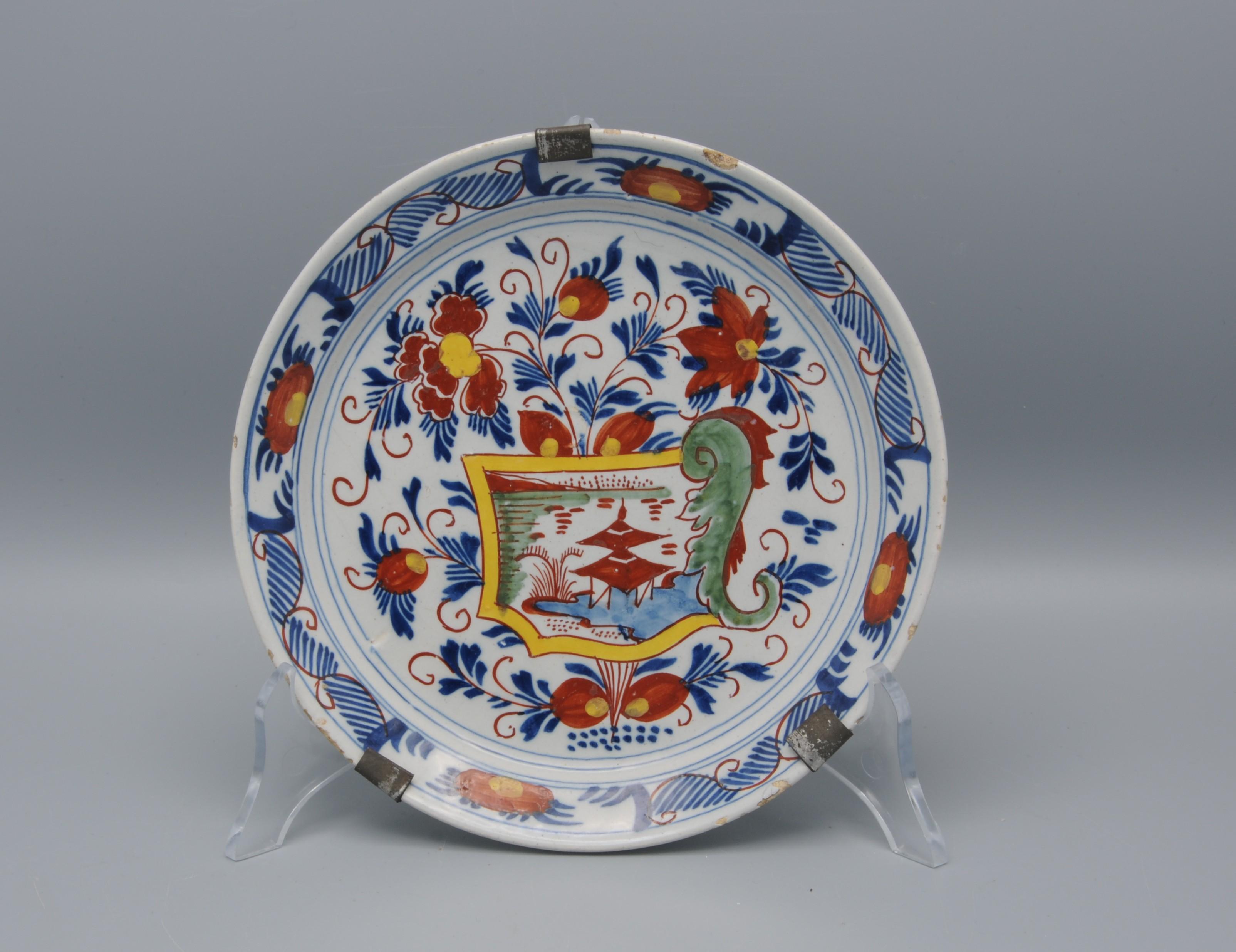 Delfter Porzellanteller aus dem frühen 19. Jahrhundert mit polychromem Kartuschen-Dekor, das eine orientalische Landschaft darstellt, die von Blumen mit Blattranken in Gelb, Blau, Grün und Eisenrot geschmückt und umgeben ist. 
Seltene kleinere