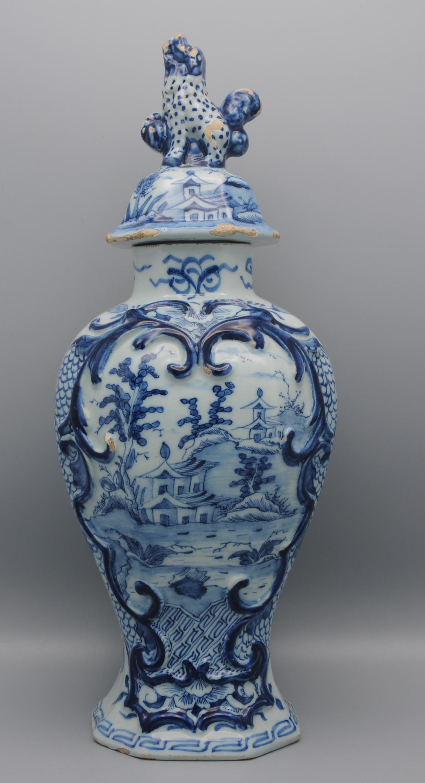Jarrón con tapa de Delftware azul de finales del siglo XVIII con decoración central de un paisaje chinoiserie con casas orientales dentro de una cartela rococó en relieve. 
Remate con decoración similar y montado con una criatura parecida a un perro