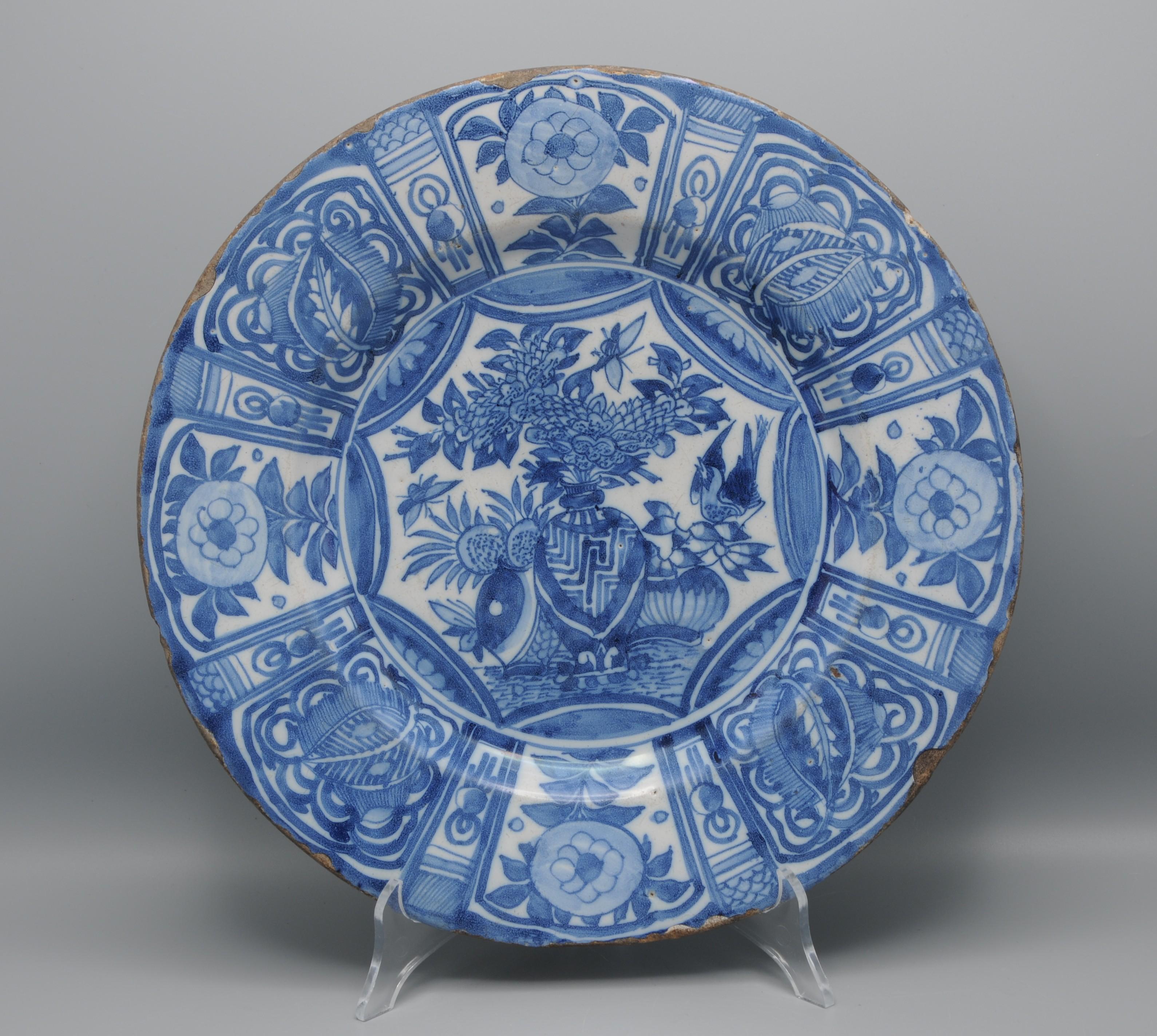 Fine Delftware dish painted in cobalt blue on tin-glazed faience, featuring central vases filled with blossoming branches, accompanied by a bird and insects. The border is divided into eight large panels alternating with smaller panels: four of the