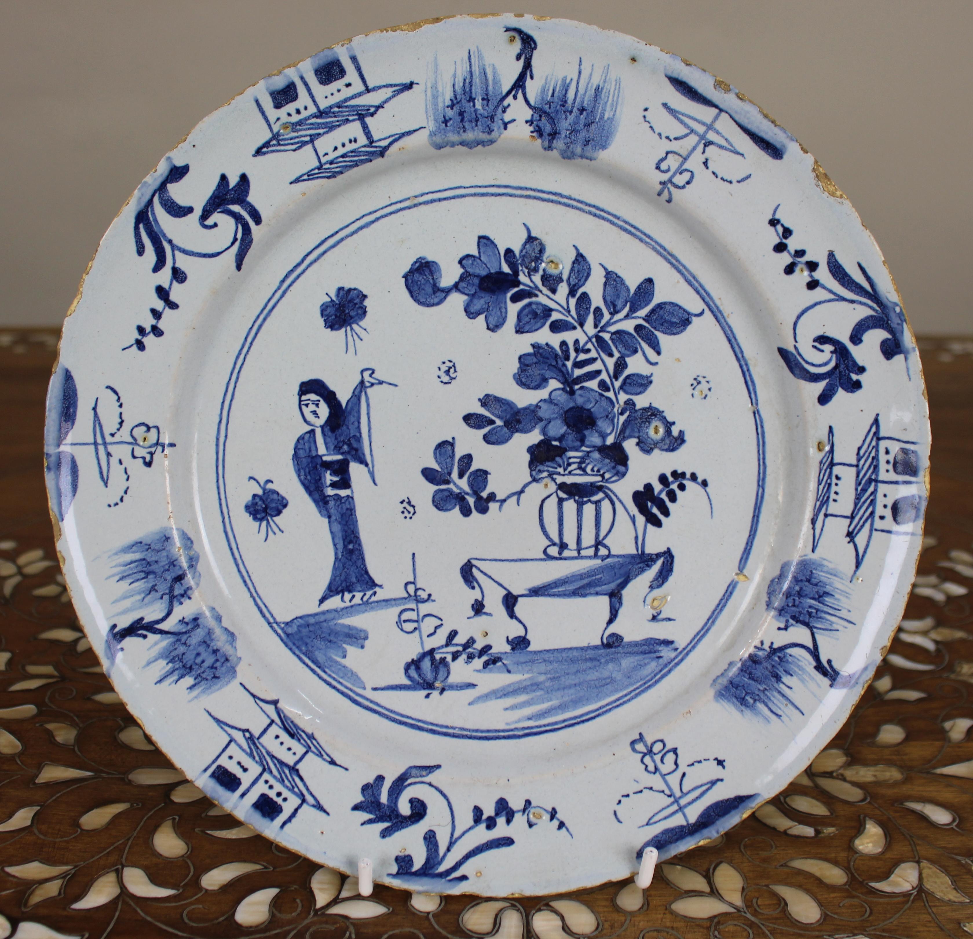 Delfter Chinoiserie-Teller, um 1760

Ein Delfter Teller aus der Mitte des 18. Jahrhunderts, blau bemalt mit einer lebhaften Chinoiserie-Szene. Das zentrale Motiv zeigt eine gewandete orientalische Figur, die eine Fahne hält und neben einer