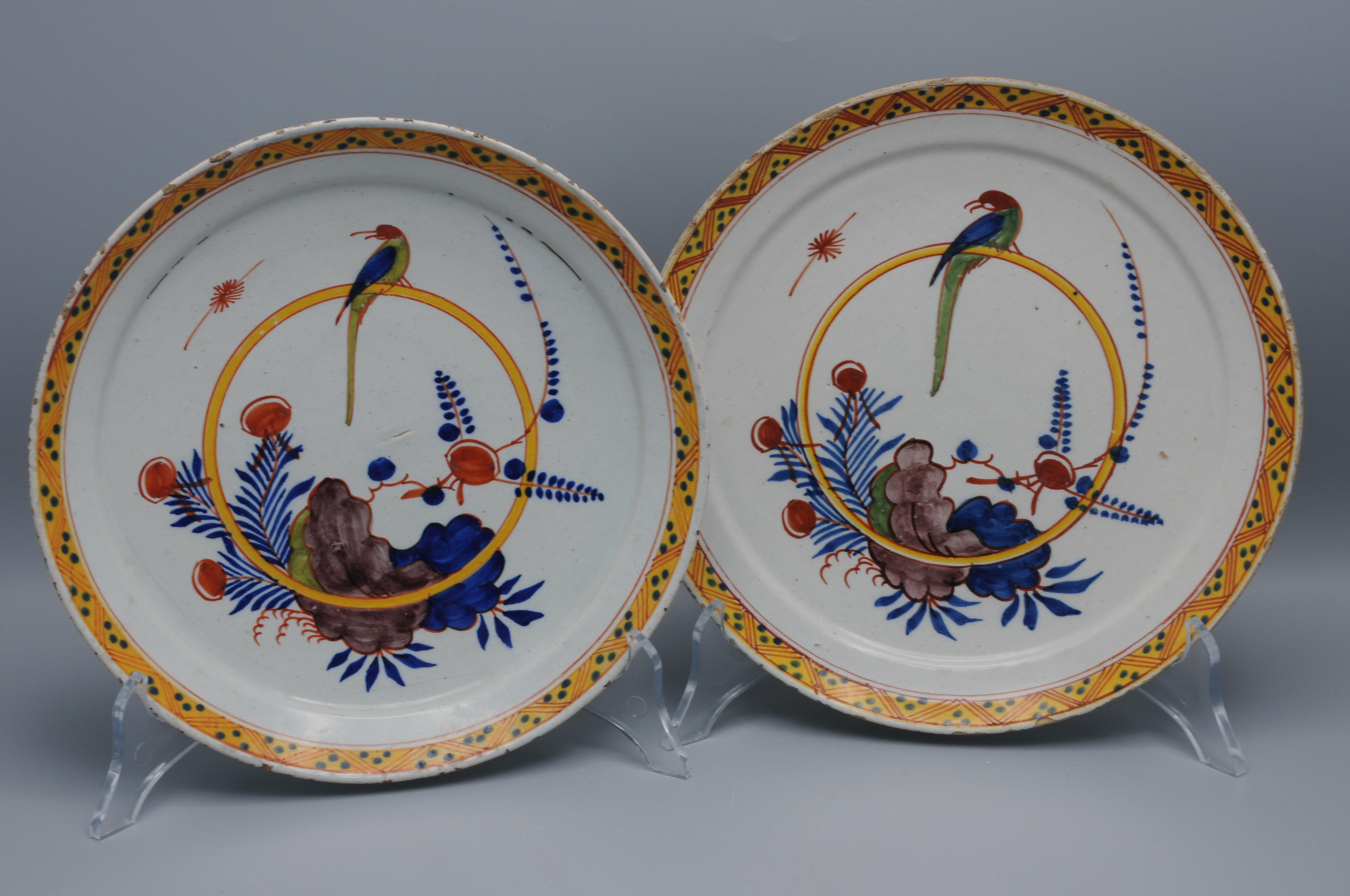 Un par de platos de Delftware policromado con un loro posado en una anilla en el centro, con interesantes elementos de paisaje asimétrico que muestran una base en el diseño de principios del siglo XVIII. Destacan la parquedad de la composición y el