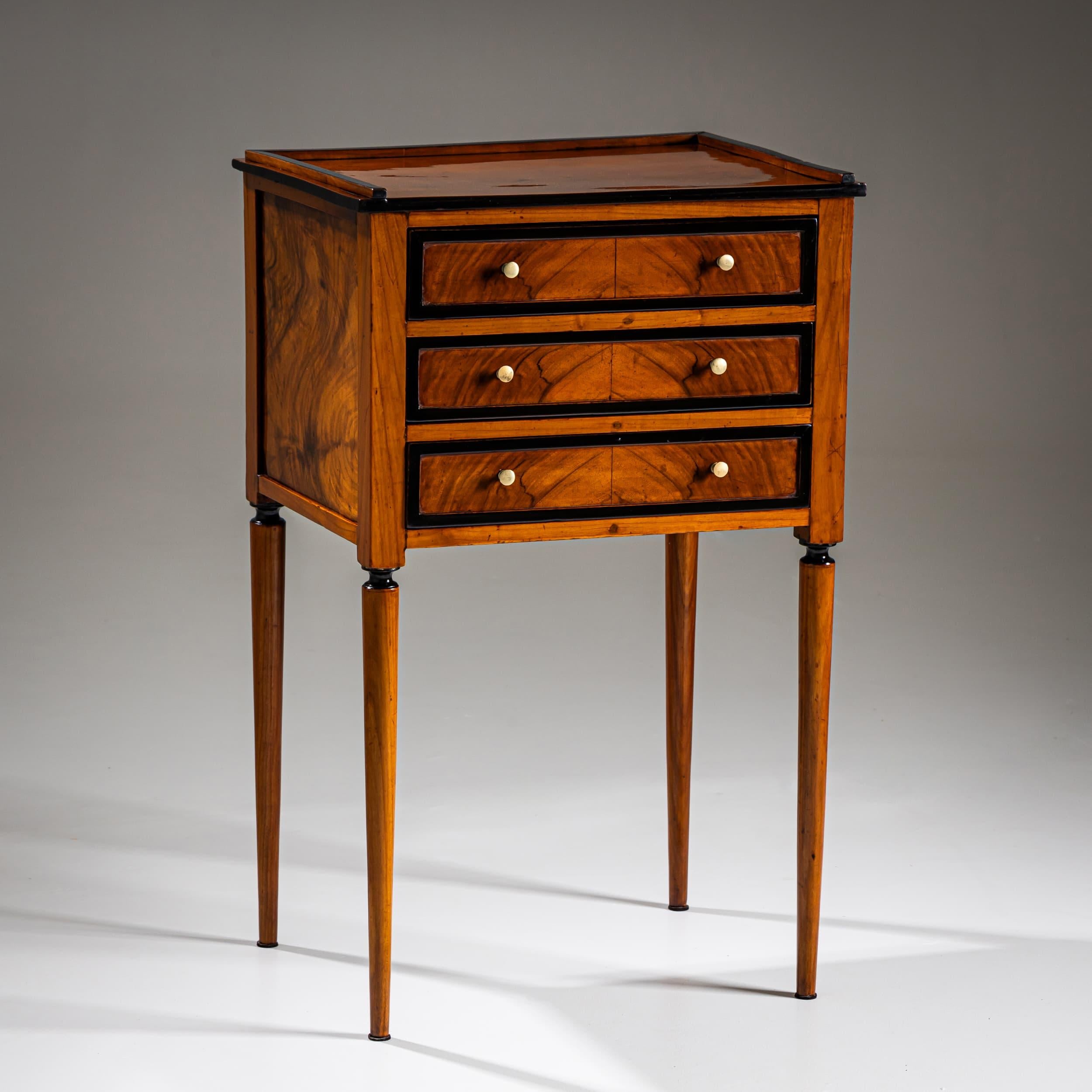 Finely crafted small commode with three drawers framed by ebonized accents. The piece stands on tall, tapered legs and features a low gallery rail along three sides of the top. Veneered in beautifully grained wood, the drawer fronts display a