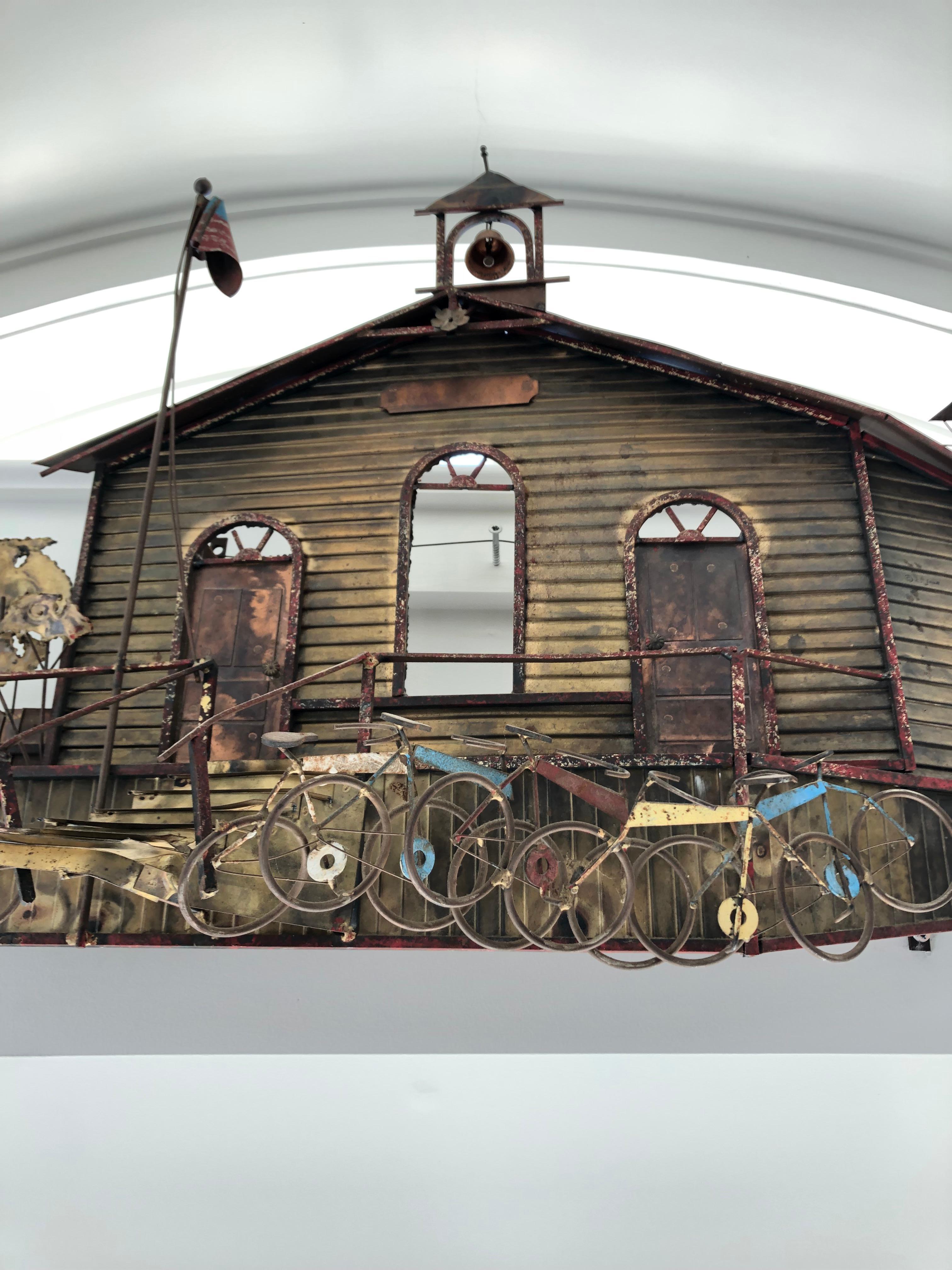 A large meticulously crafted 3 dimensional wall sculpture made of metal and brass of a school with bell tower, arched windows and door, bicycles parked in front and an American flag. The more you look, the more ingenious details are