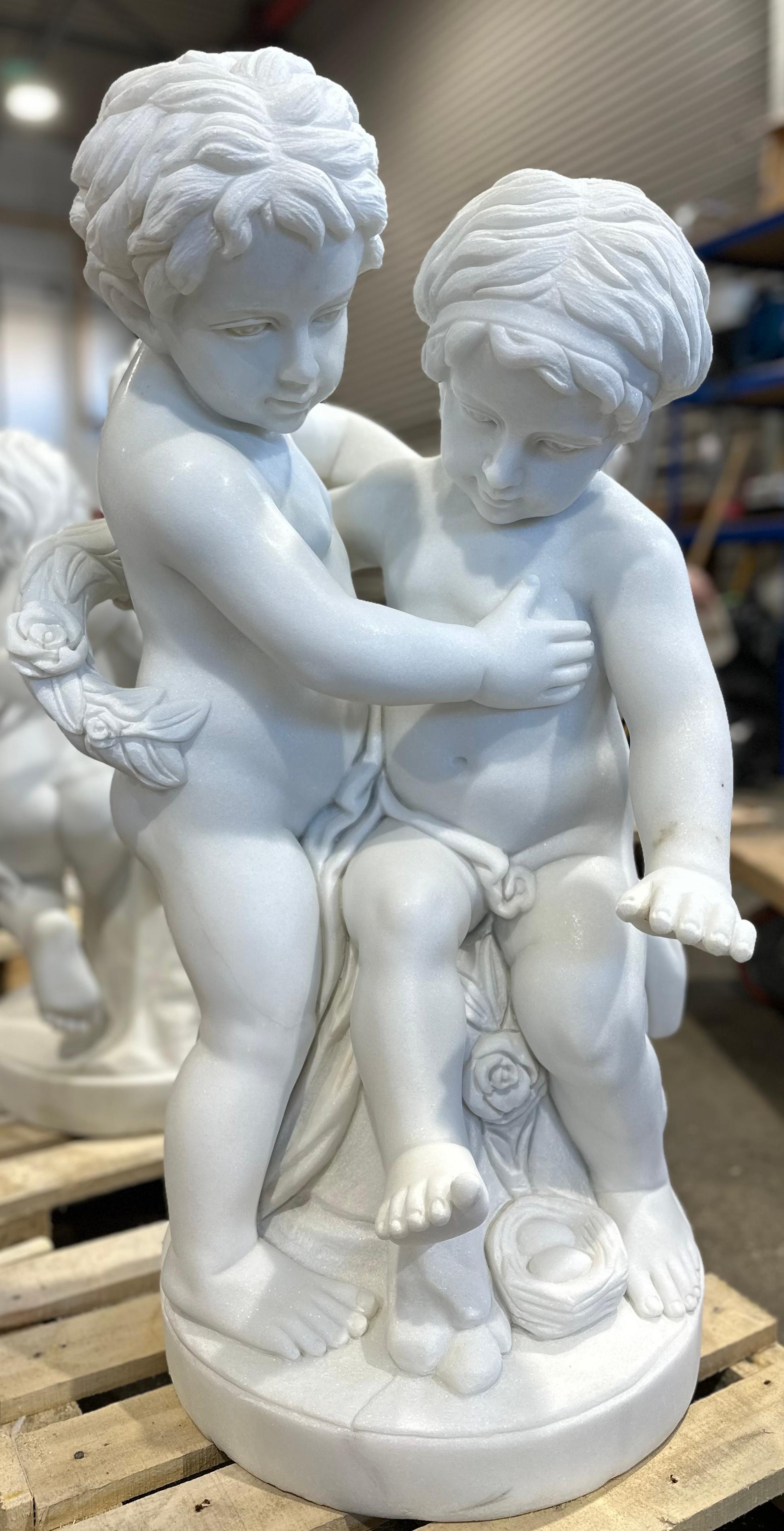 A enchanting white marble sculpture of two childlike putti, one standing the other seated, with a floral wreath wrapped around them. And a birds nest with two eggs at their feet. 
The carving is well done and detailed from the ruffled hair of the