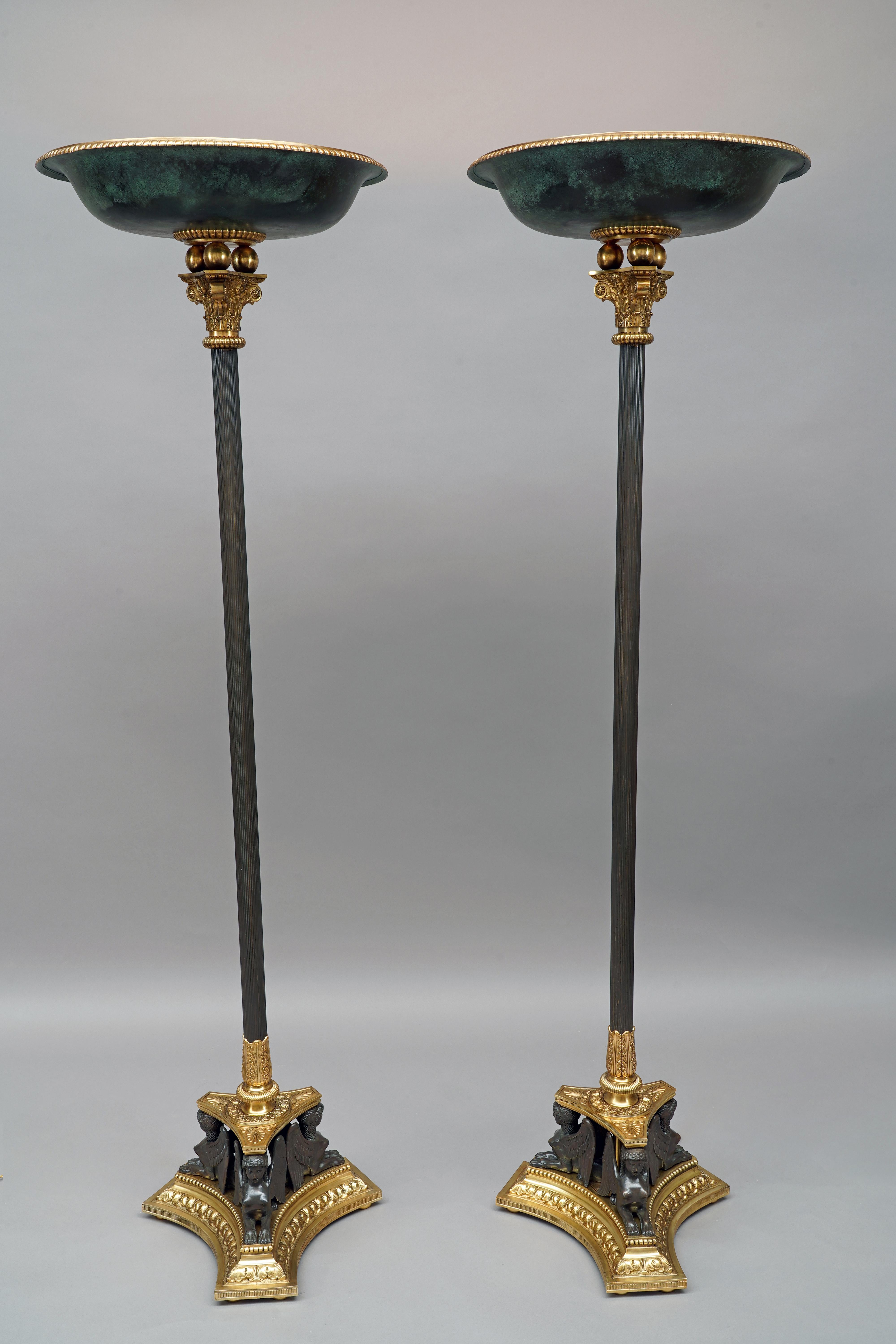 Coppia di lampade da terra di ispirazione antica in metallo e bronzo patinato e dorato attribuite a Delisle. Il fusto scanalato è sormontato da un capitello dorato decorato con foglie d'acanto e una testa di cherubino alata, e da tre sfere che