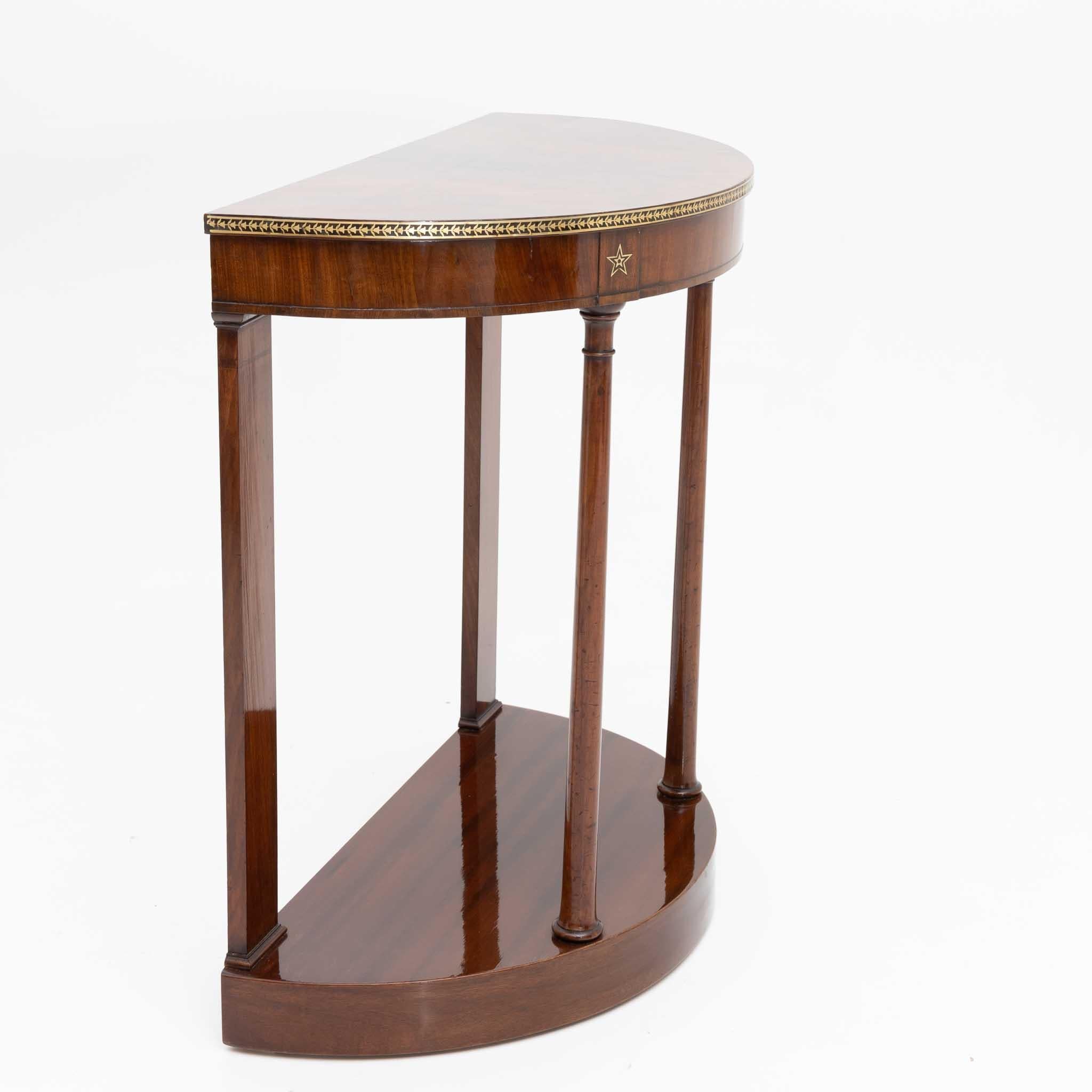 Demi Lune console veneered in mahogany with pilasters and conical column decor, smooth plinth zone and frame with brass inlays in the form of stars and floral motifs.