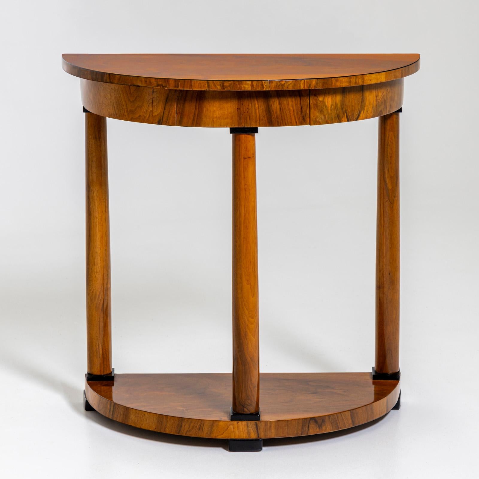 A Biedermeier-style console table featuring a demi-lune tabletop and base supported by three columns. The sleek frame includes a slim, built-in drawer. The table is veneered in walnut and has been hand-polished.