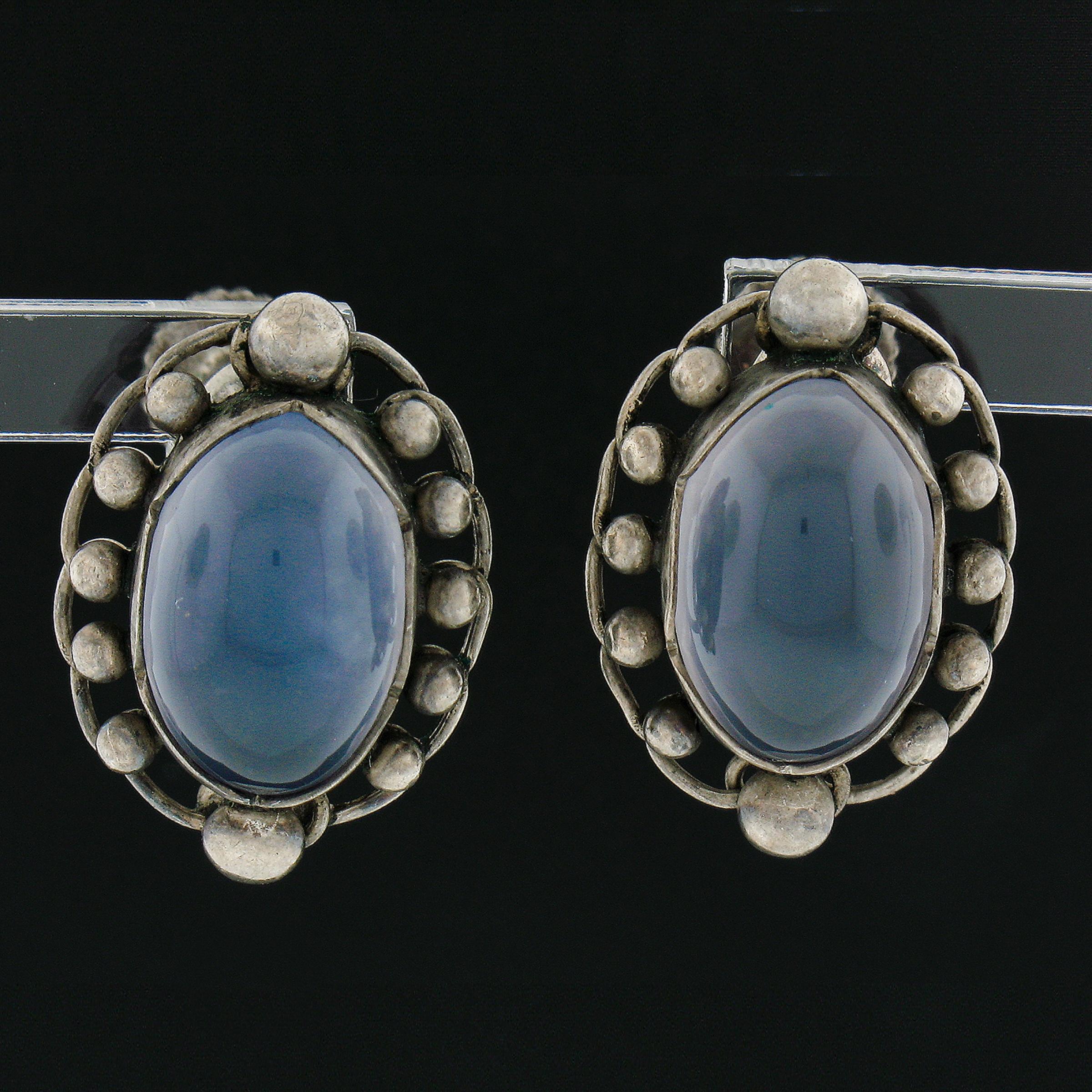 These adorable, vintage made in Denmark #81 pair of earrings By Georg Jensen are crafted in solid sterling silver and feature bezel set chalcedony. The frame is beaded open work and hand made. These earrings are secured with their original screw-on