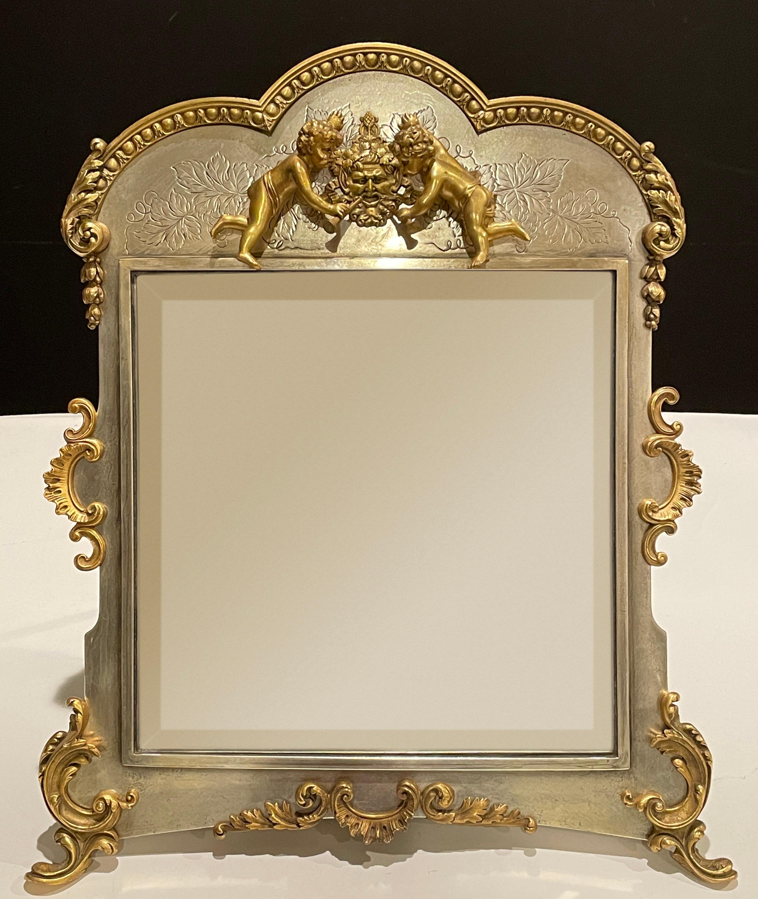 An American Silver-Plate and gilt table mirror 19th century. 
In the Rococo taste, the floral and foliate scroll decorated frame surmounted by a putto with Bacchus mask blowing trumpets raised on scroll feet, marked Derby Silver Co. Derby Conn. on