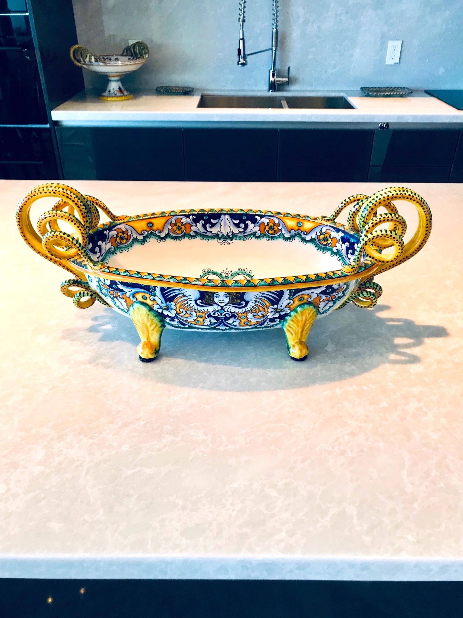 Exquisite Majolika-Schale mit handgemalter Zinnglasur auf Steingut-Keramik. Handgefertigt mit stilisierten:: schlangenförmigen Griffen und aufwendig bemalten Mustern. Die Schale hat eine ovale Form mit einem Sockel. In leuchtenden Gelb-:: Blau- und