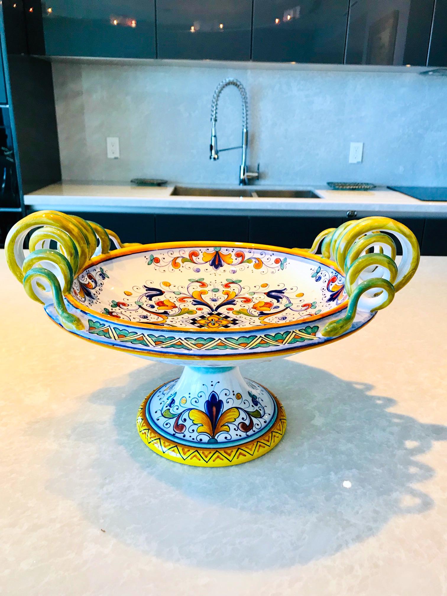 Exquisite Majolica centerpiece bowl with hand painted tin glaze over earthenware pottery. Handmade with pedestal base and stylized serpentine handles and elaborate painted designs throughout. In vibrant hues of yellow, blue, and green over white.